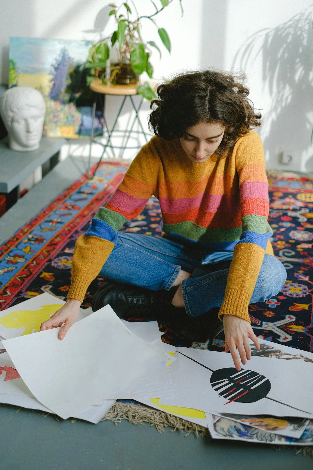 Person sitting cross-legged on a patterned rug, wearing a colorful striped sweater, examining artwork scattered on the floor.