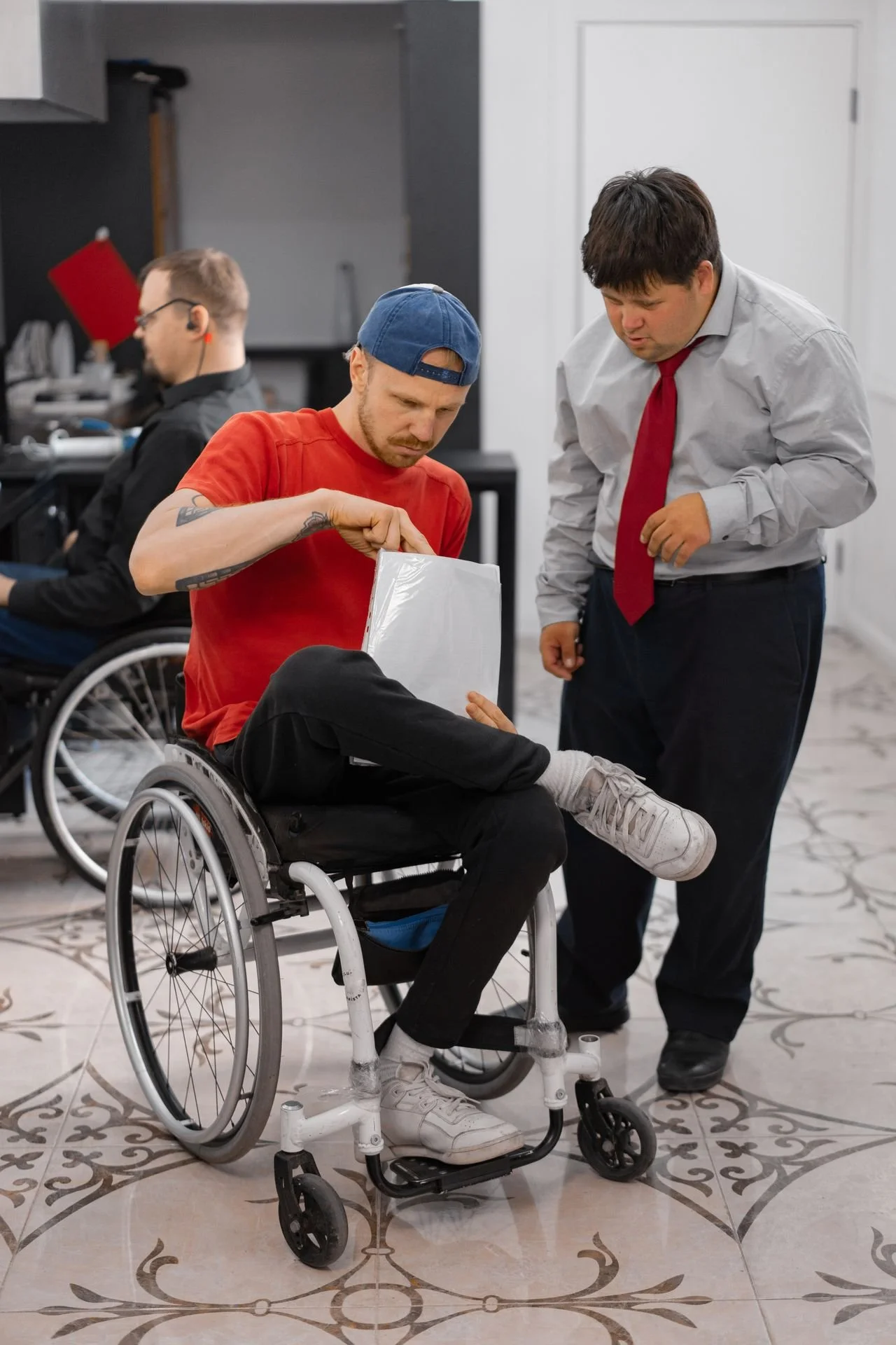 Man in wheelchair wearing a red shirt and blue cap looks at a document while a man in a suit with a red tie stands beside him indoors.