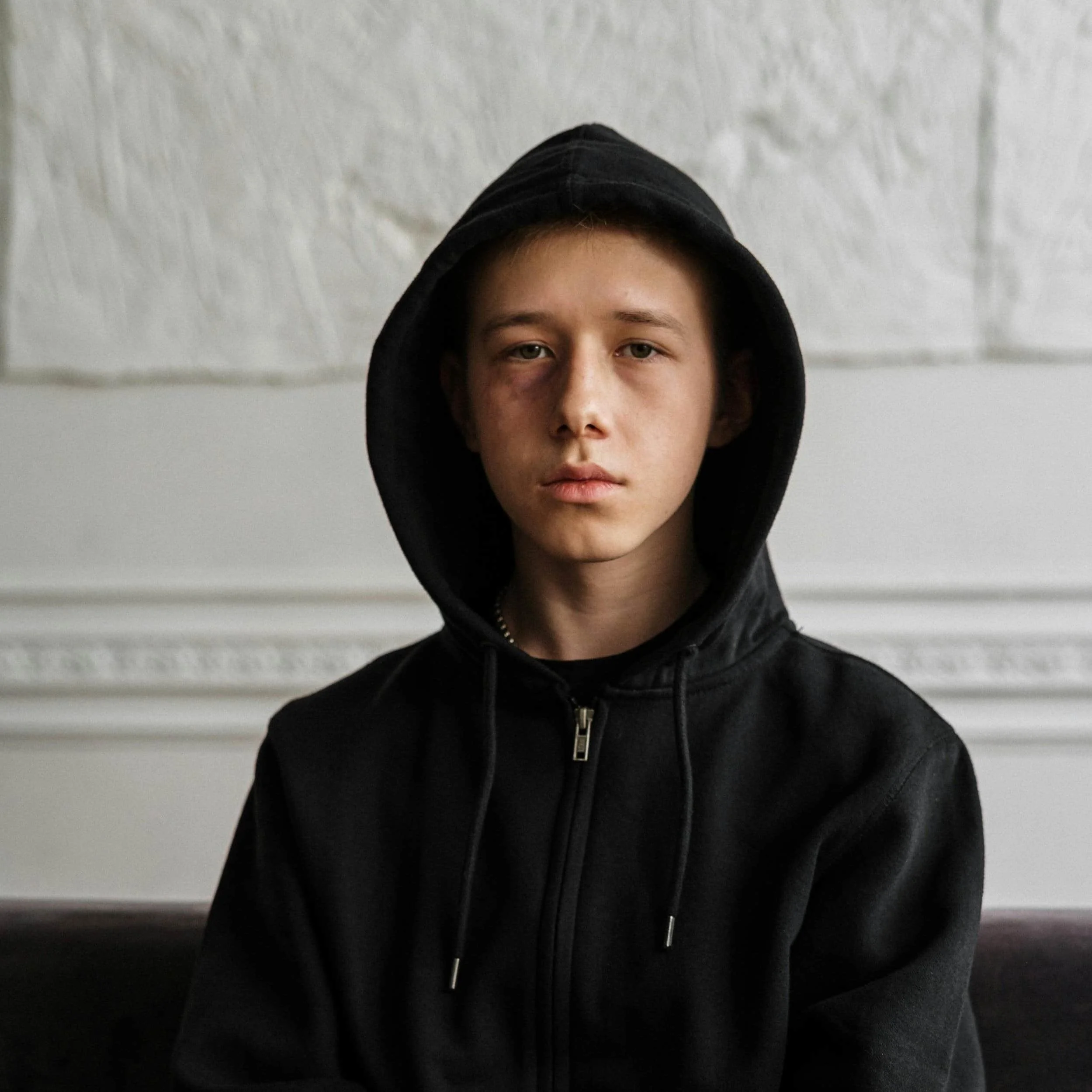A person wearing a black hoodie sits on a couch, against a textured white wall, exuding a sense of mystery and contemplation.