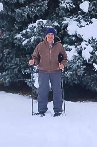A person stands in a snowy landscape, holding ski poles, dressed warmly with a brown jacket against a backdrop of evergreen trees.