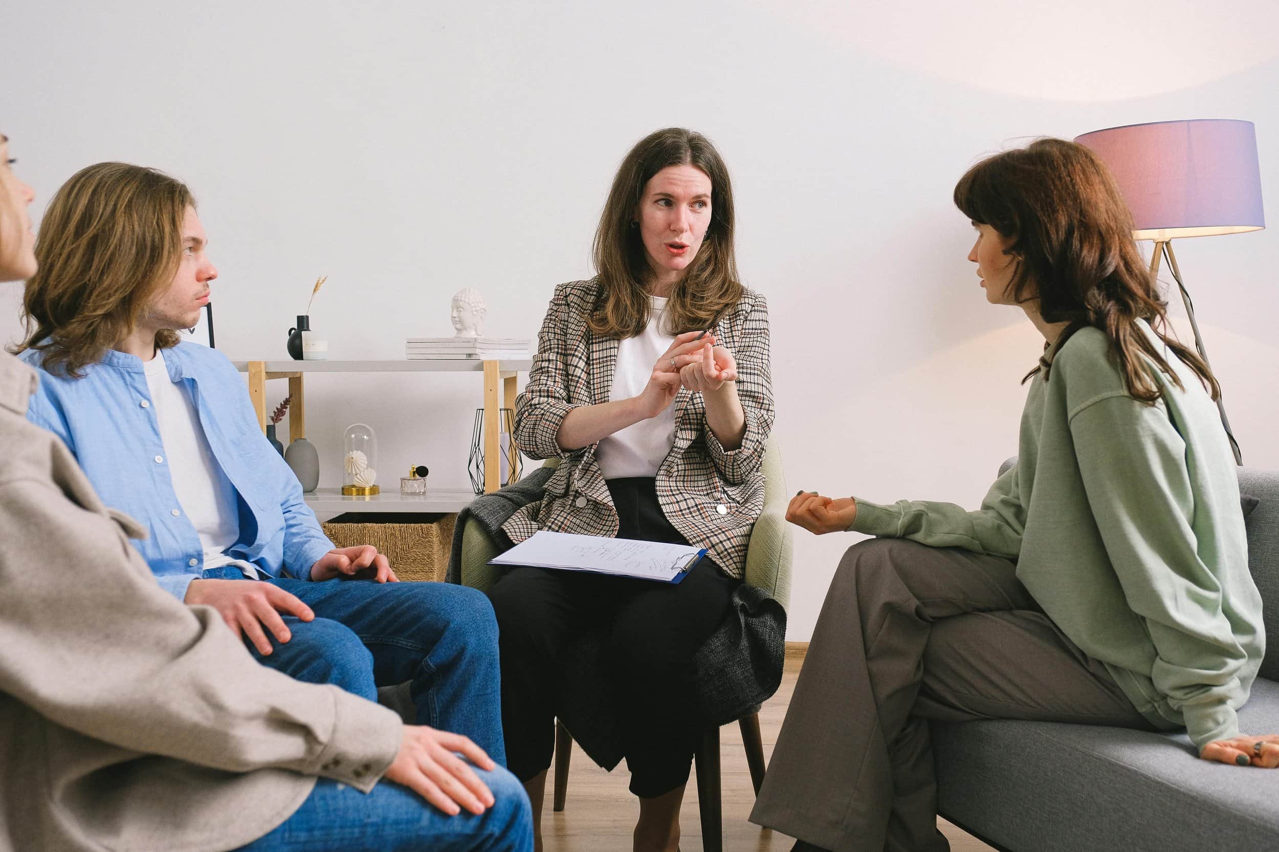 Four people in a therapy session discussing, seated in a bright, cozy room with shelves and a lamp in the background.