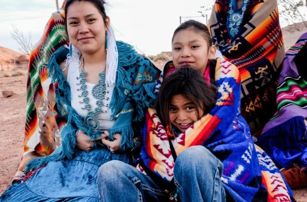 "A Native American family dressed in traditional clothing sitting together outside smiling."