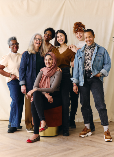 A group of 7 women of various ethnicities and cultures stand together smiling and enjoying connection