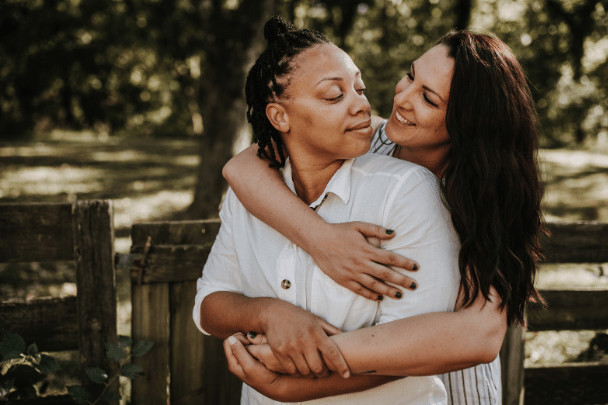 Two women embrace in a sunlit outdoor setting, surrounded by trees and a wooden fence, radiating warmth and affection.