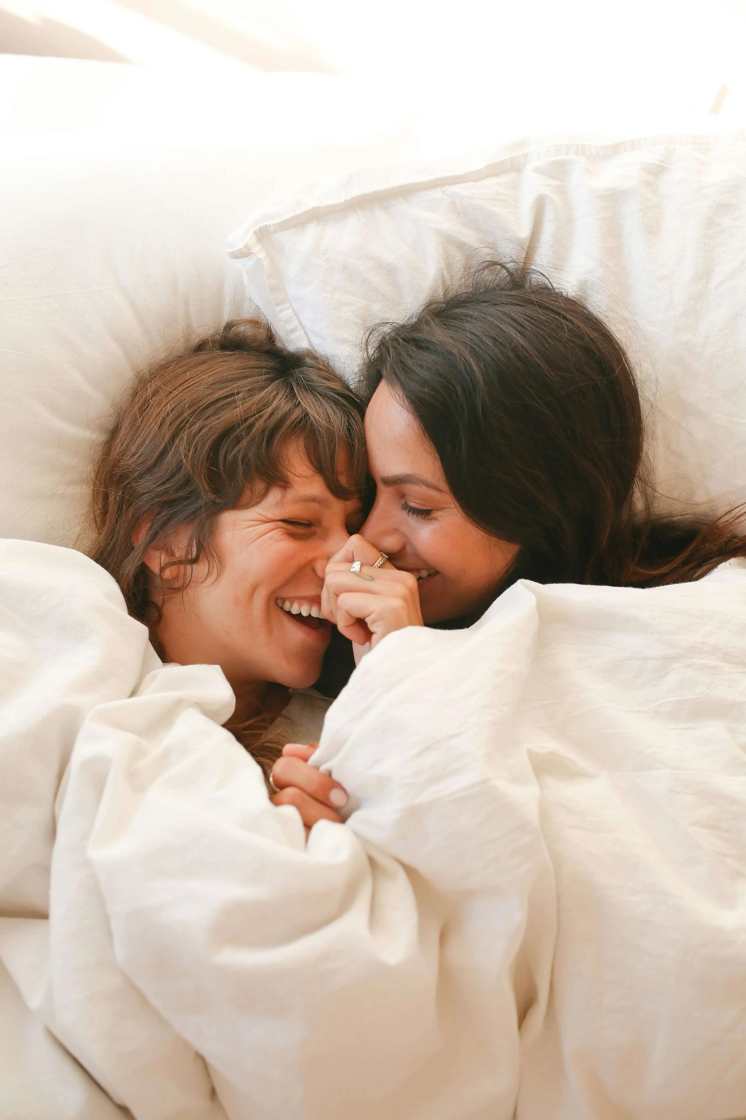 Two people lying in bed, snuggled under white blankets and pillows, facing each other closely.