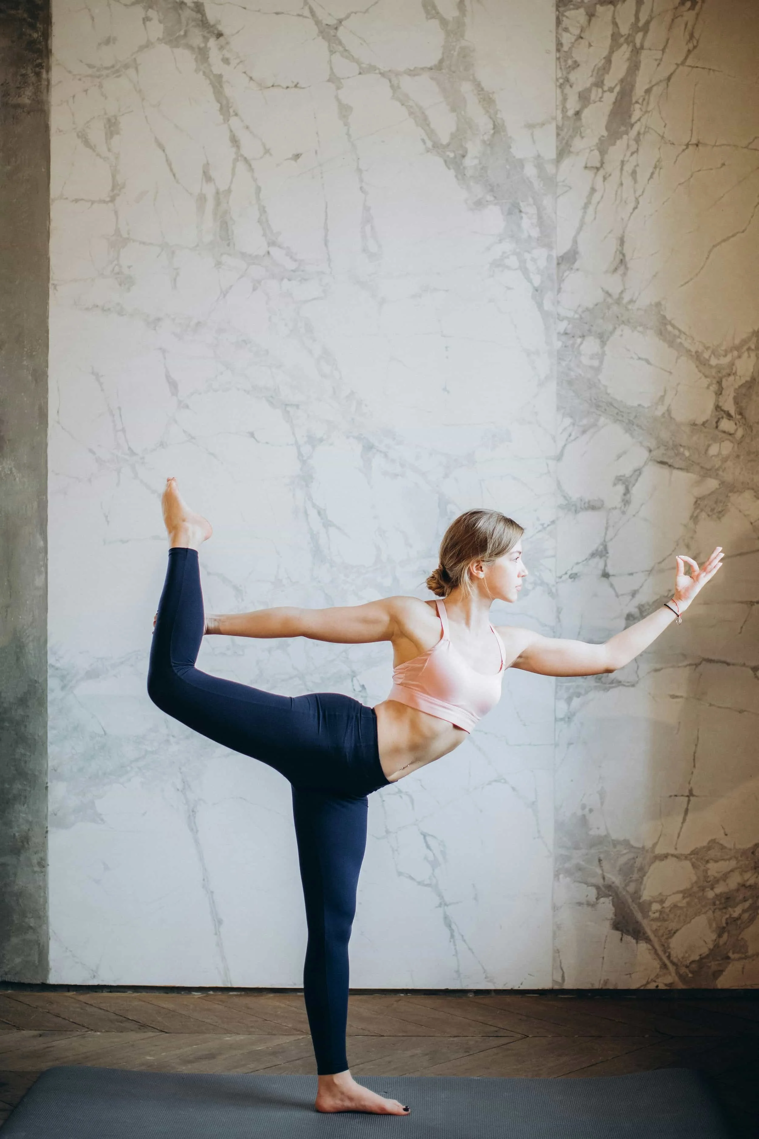 Woman in pink sports bra and black leggings balancing on one leg in yoga pose indoors on a mat against a marble wall background.