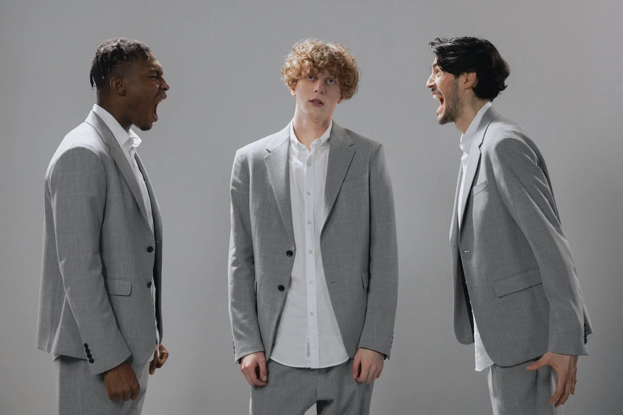 Three men wearing gray suits and white shirts stand against a plain gray background, facing different directions two are yelling at each other and one stands in the middle rolling his eyes