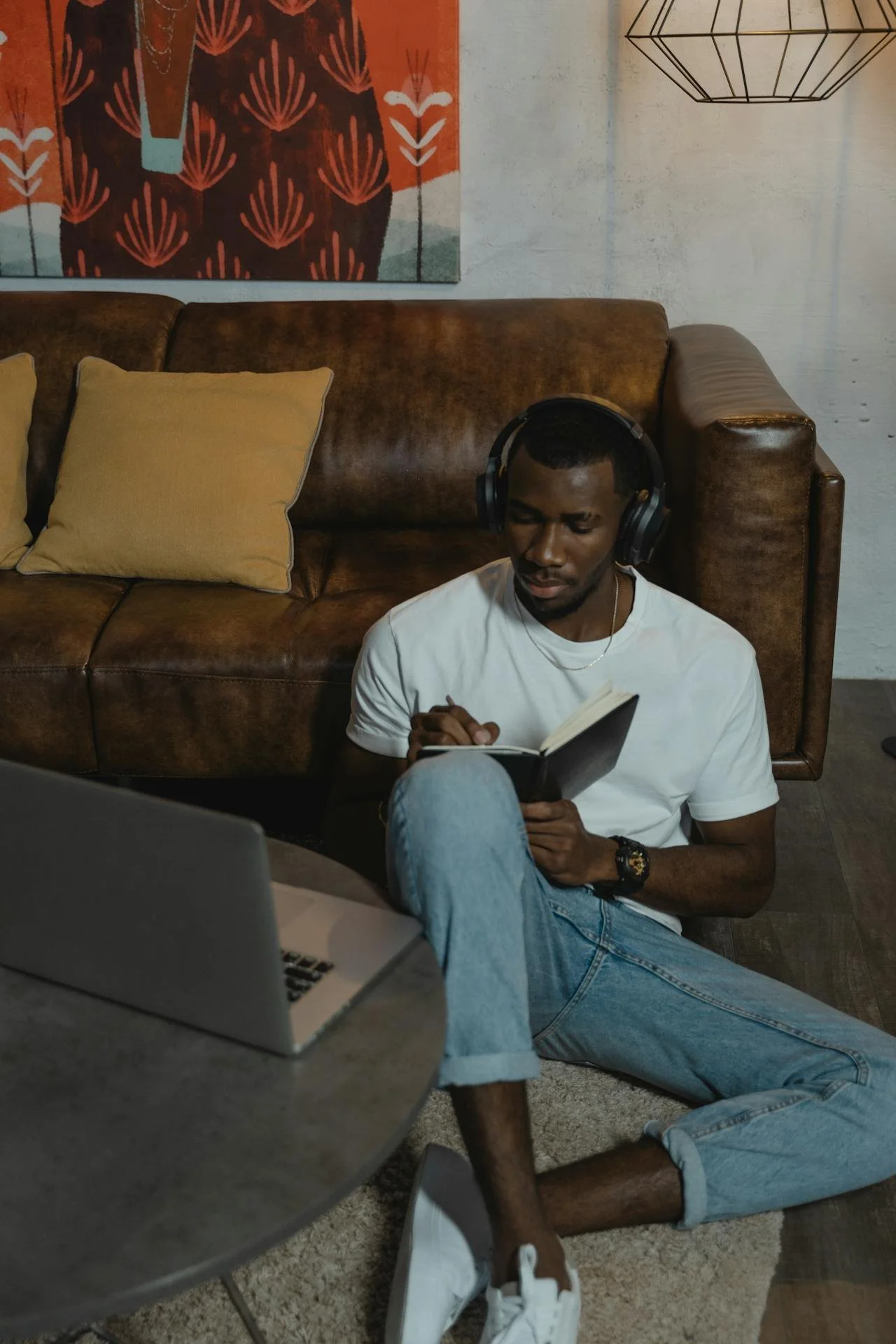 Person sitting on the floor with headphones, writing in a notebook, next to a laptop and a brown leather couch with pillows.