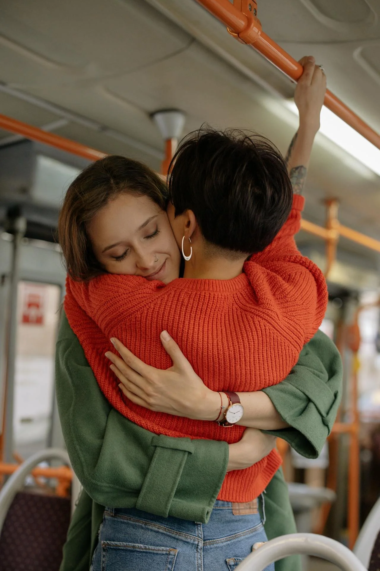 Two women hugging on a bus, one wearing a red sweater and the other wearing a green coat.