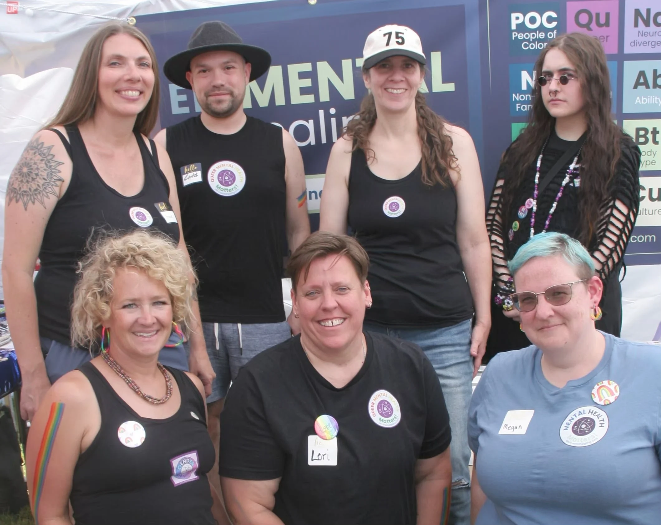 Seven people pose together outdoors, smiling, wearing black or blue shirts with pride pins and badges, with a diversity banner behind them.