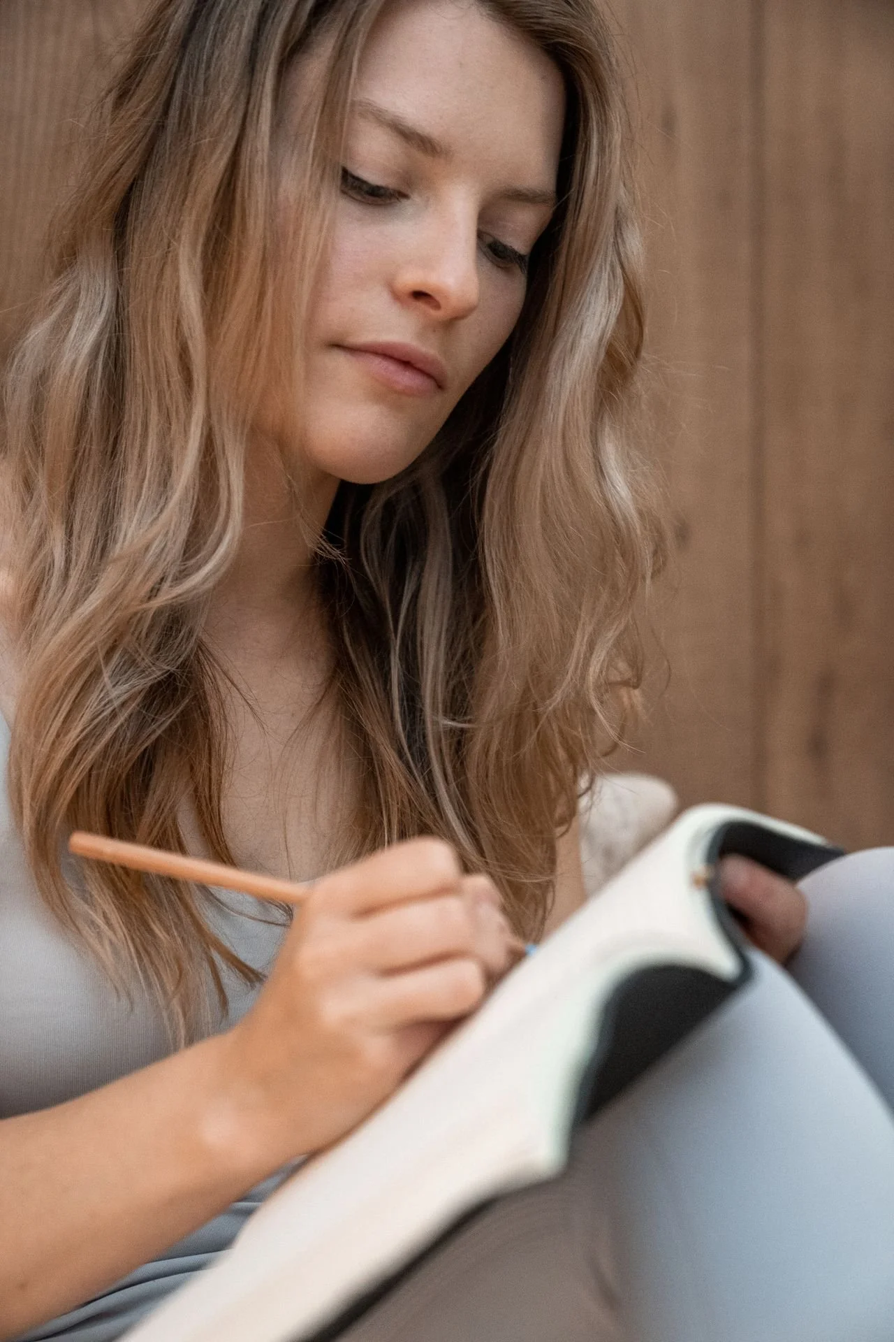 Woman with long blonde hair writes in a notebook with a pencil, sitting indoors against a wooden background.