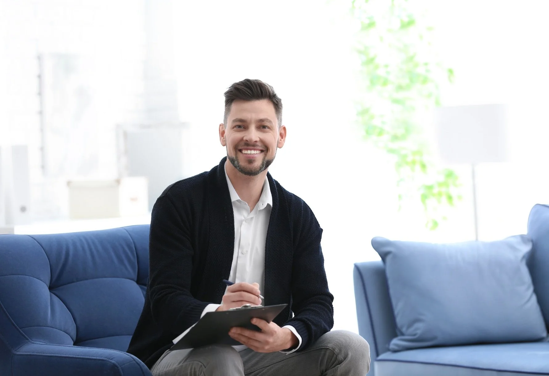 Man sitting on a blue couch holding a clipboard and pen, dressed in a white shirt and dark cardigan in a bright room.