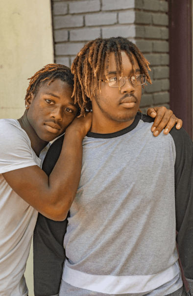 Two people with dreadlocks standing close, one resting head on the other's shoulder, showing a moment of comfort and support.
