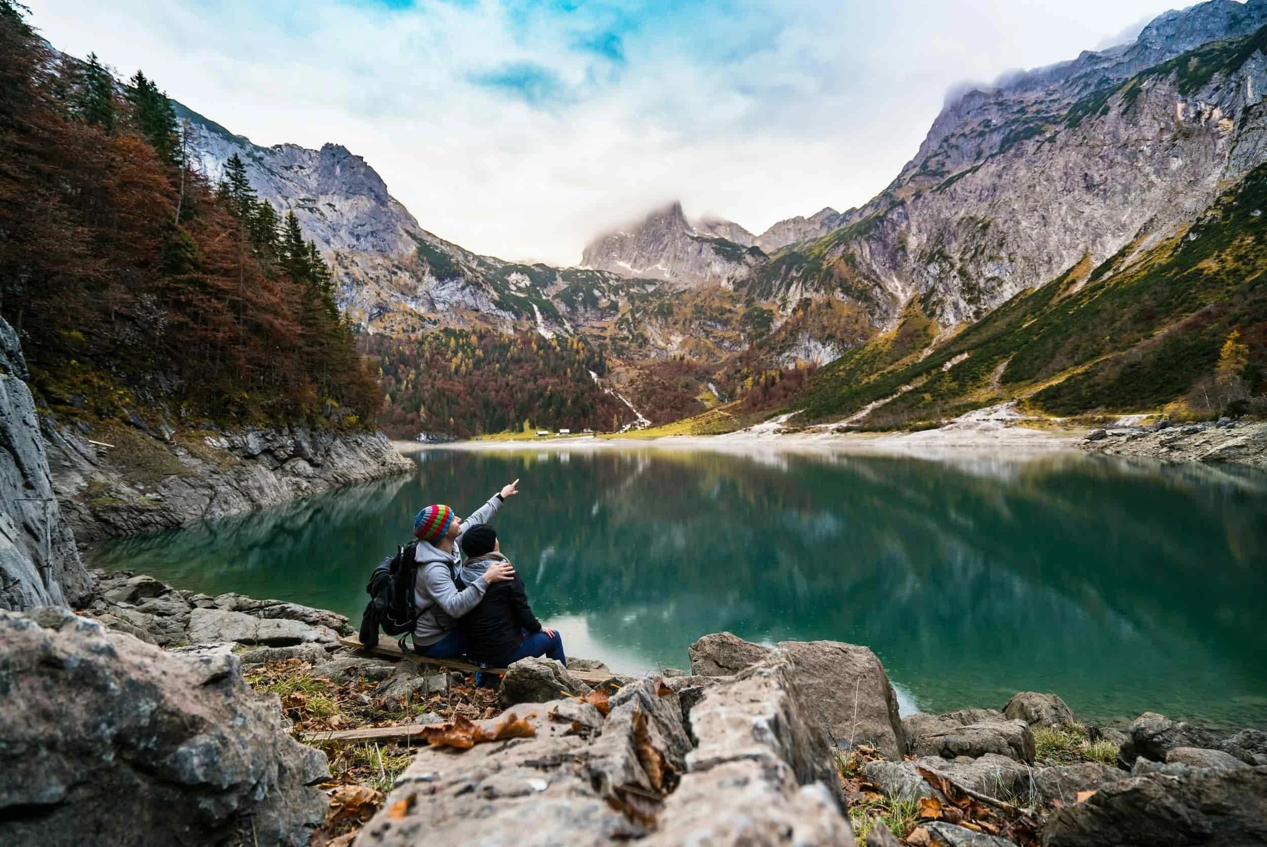 A couple sits on rocks by a serene lake, pointing at the mountains, surrounded by autumn foliage and a clear blue sky.