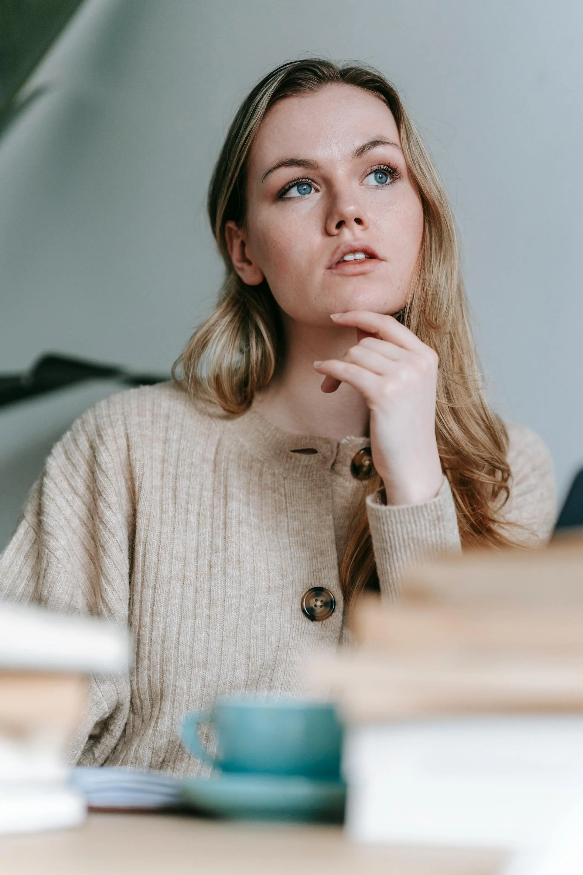 Person in a beige sweater sitting at a table with books and a green coffee cup, resting their hand on their chin.