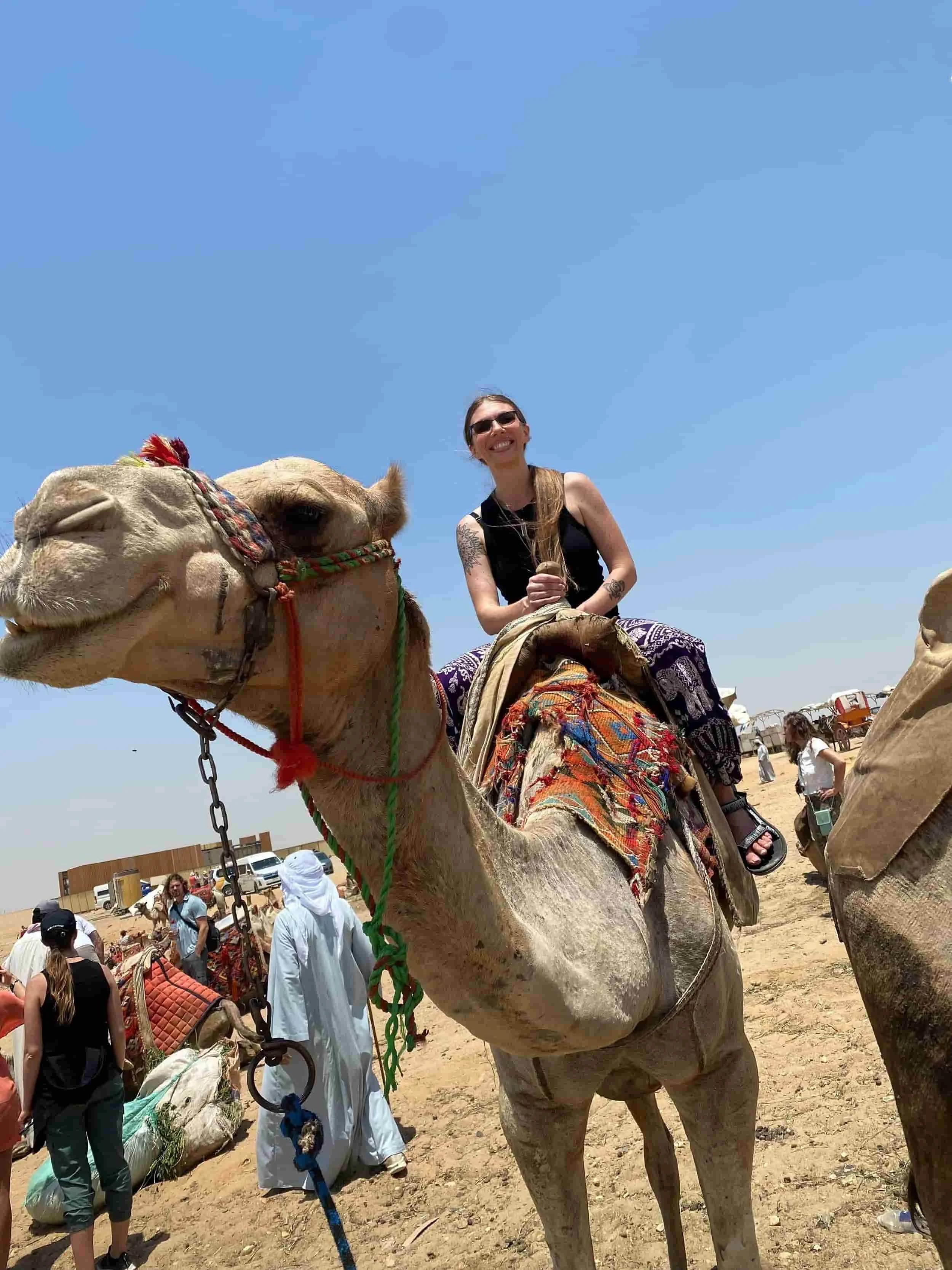 Person riding a decorated camel in a desert setting with others and tents in the background under a clear blue sky.