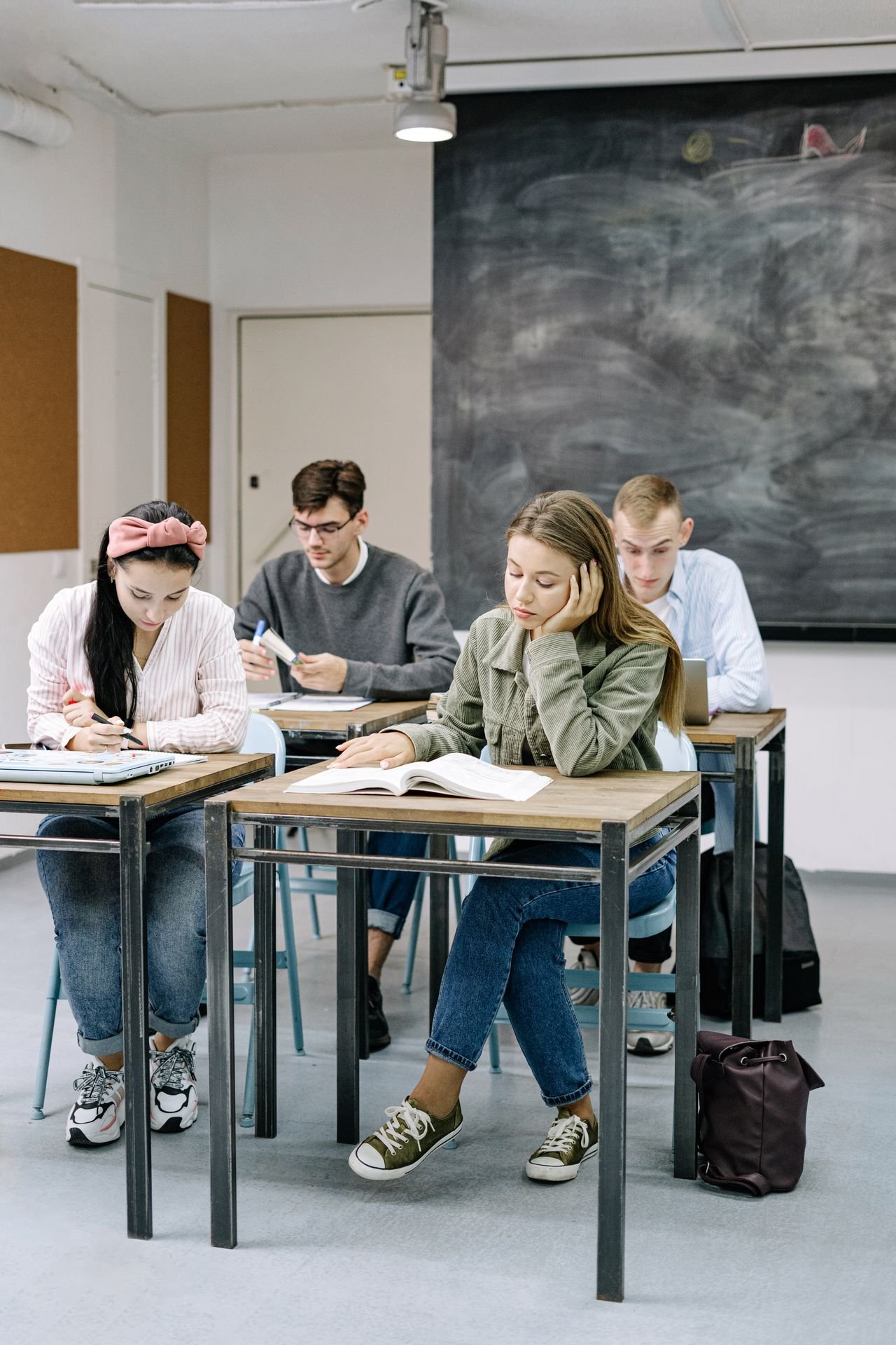 Four students sit at desks in a classroom, reading and writing, with a large chalkboard in the background.