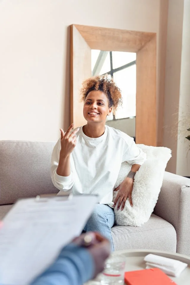 Person with curly hair sitting on a couch, gesturing while talking to someone holding a clipboard in a bright, cozy room.