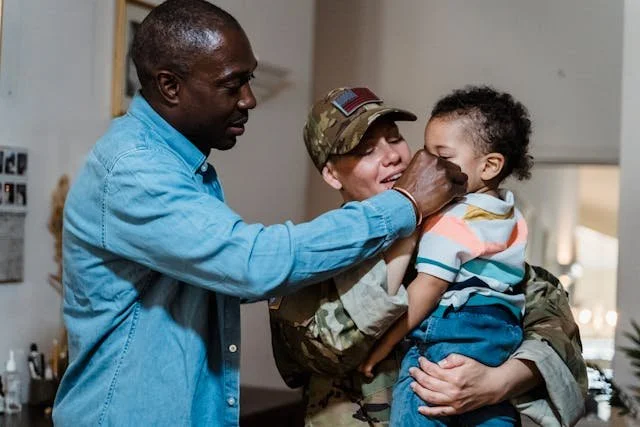 A military service member in uniform holds a young child while an adult gestures nearby, creating an emotional reunion atmosphere.