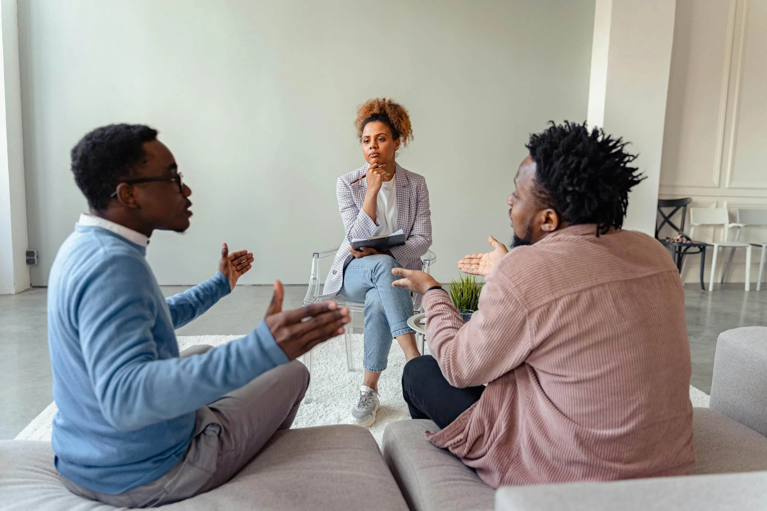 A group discussion in a modern, minimalistic space, featuring two people engaged in conversation with a seated listener holding a notebook.