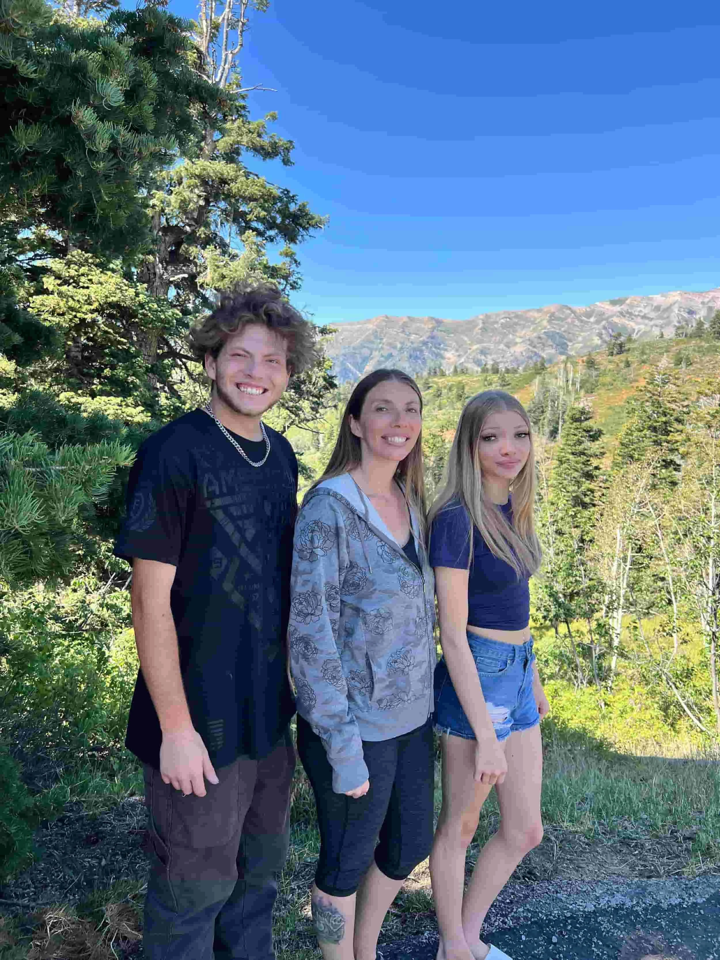 Three people standing outdoors near trees with mountains in the background under a clear blue sky.