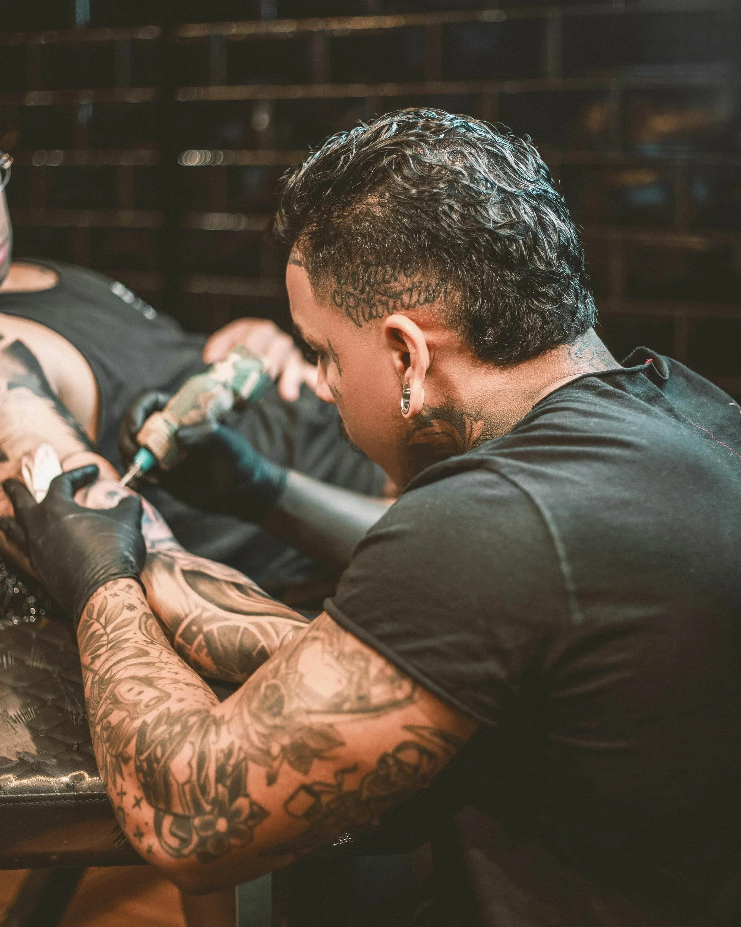 A person with tattoos on their arm getting a tattoo on their forearm in a tattoo studio, with another person working on the tattoo.
