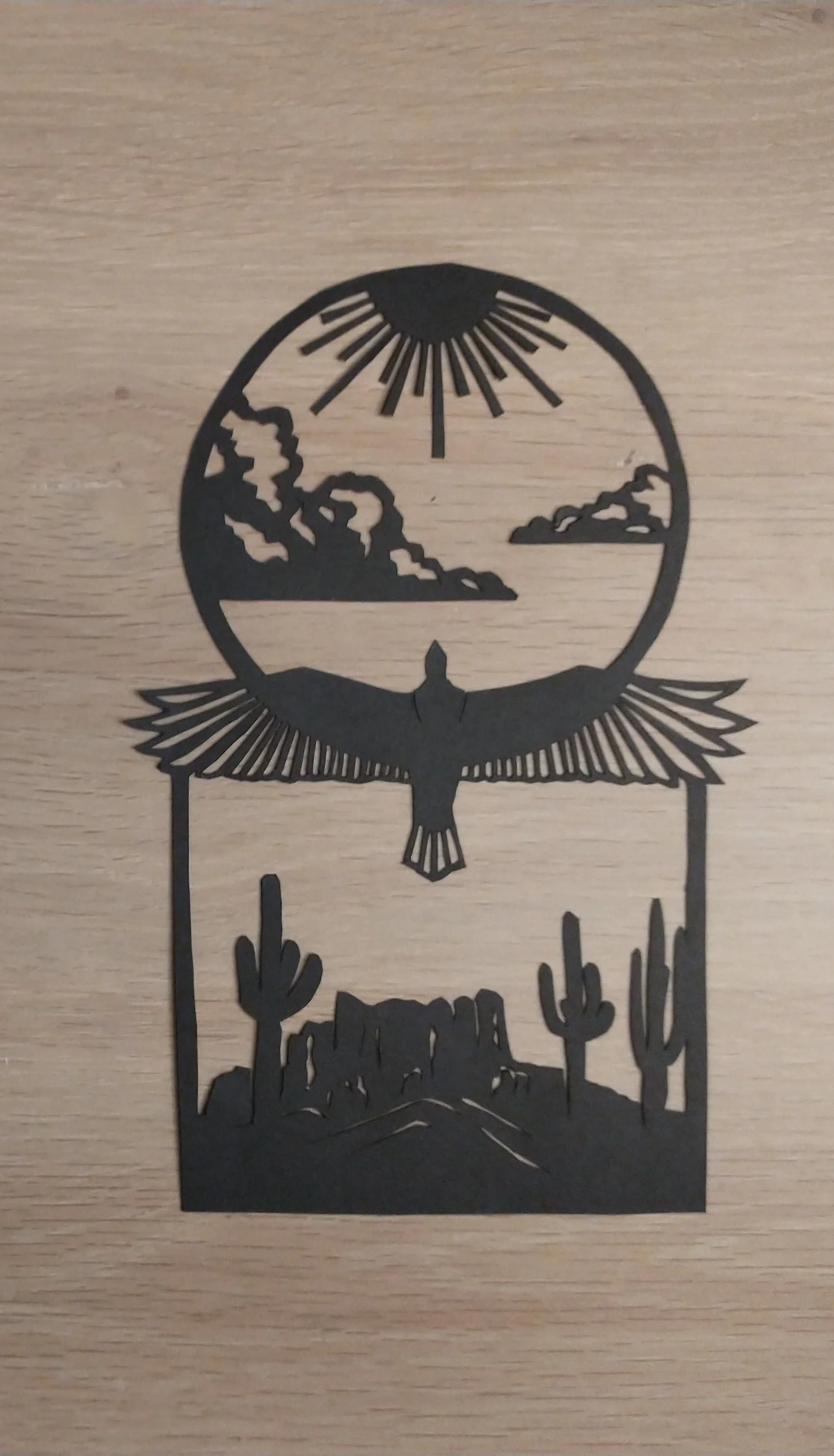 Silhouette of a desert scene with a bird flying above cactus and rocks, framed by a circle with sun rays at the top.