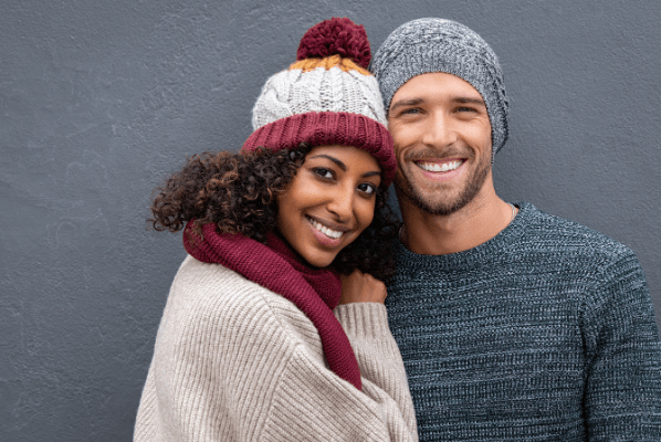 A couple stands closely together against a gray wall, both wearing cozy knit hats and warm sweaters, radiating affection.