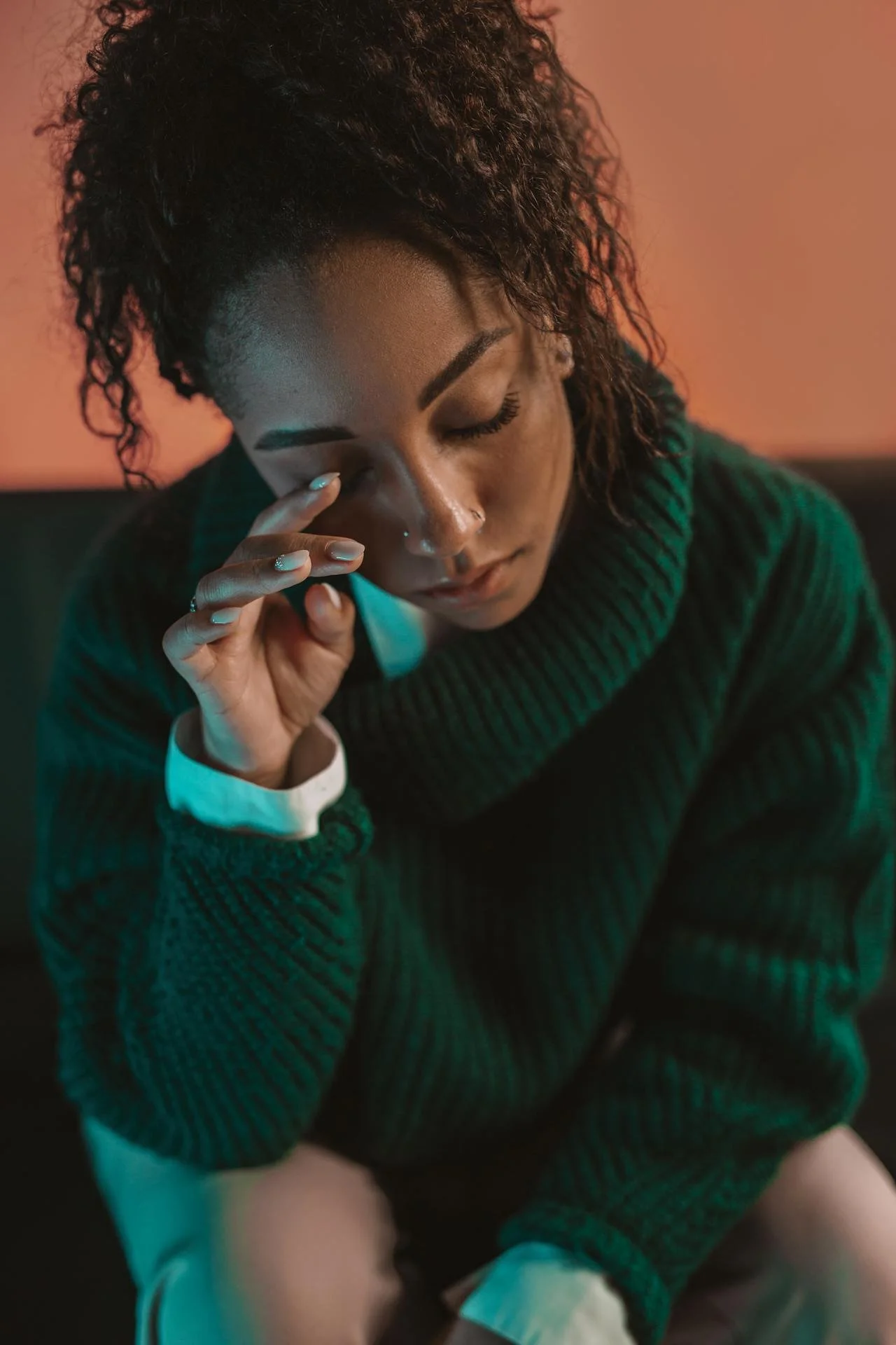 A woman with curly hair and pierced nose resting her hand on her face, wearing a green sweater, appears sad or overwhelmed.