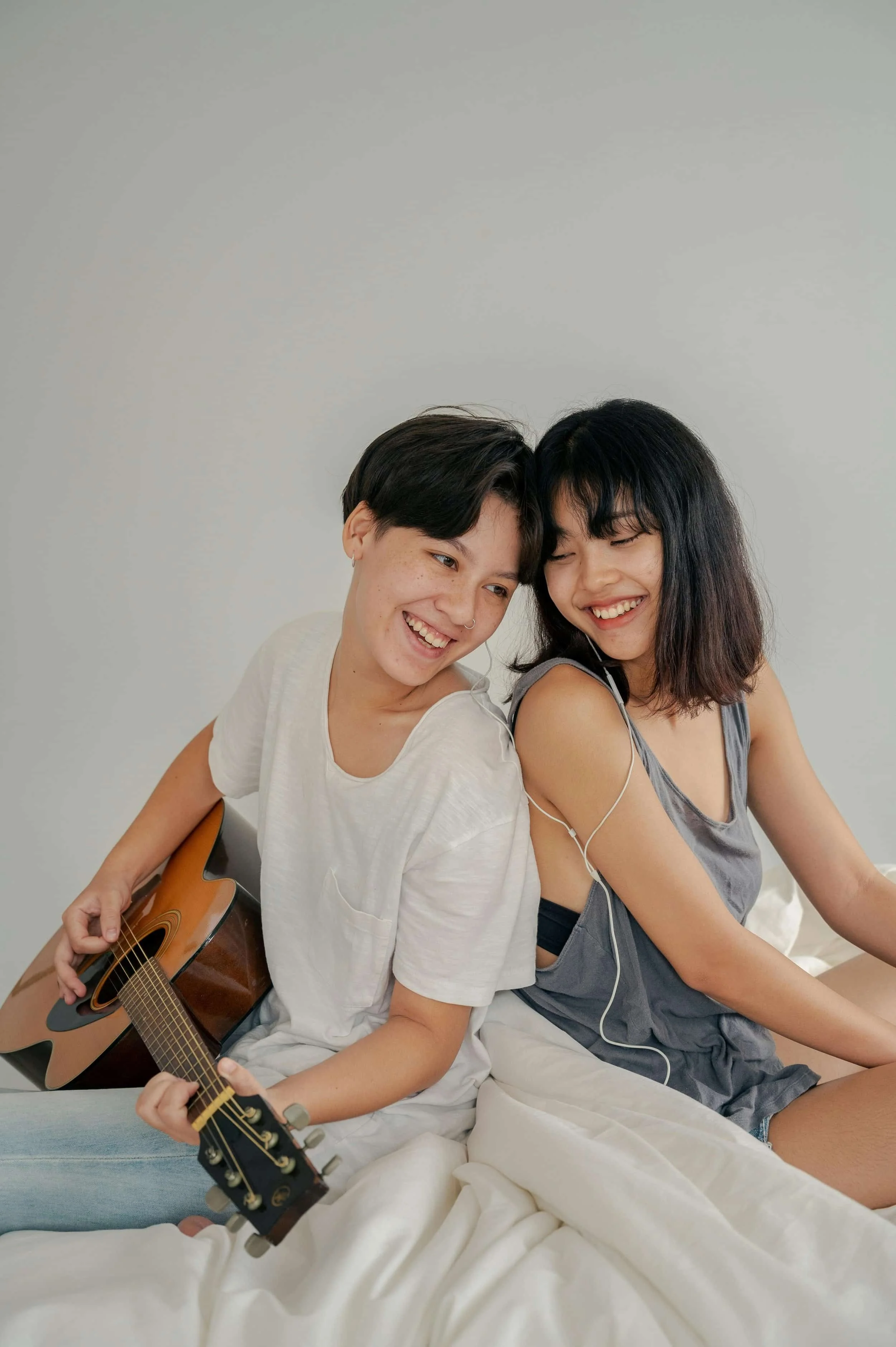 Two people sitting back-to-back on a bed, one playing an acoustic guitar while sharing earphones.
