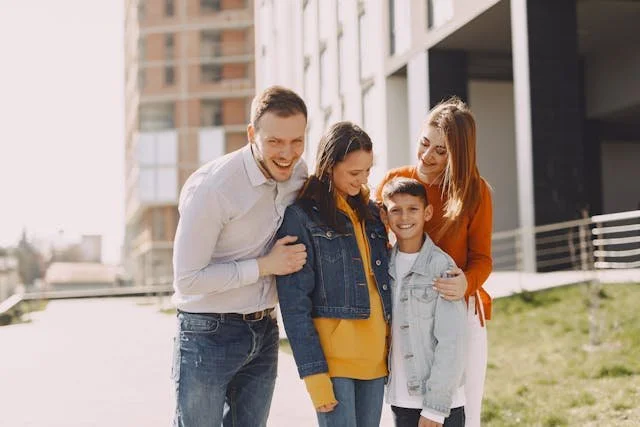 Family of four standing together outdoors near modern buildings on a sunny day, dressed casually in jackets and sweaters.