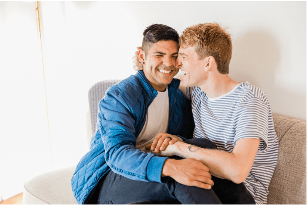 Two people sit closely together on a light gray sofa, sharing a tender moment, with hands intertwined and expressions of affection.