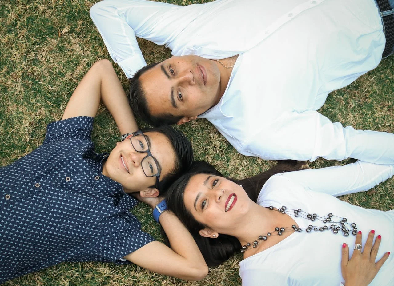 Three people lying on grass, heads together, one in a white shirt, another in a patterned blue shirt, and a woman with a necklace.