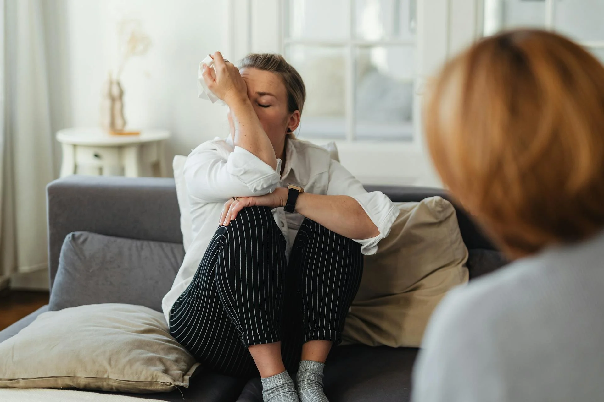 A person sits on a couch hugging their knees, wiping their eyes, while another person sits nearby, partially out of focus.
