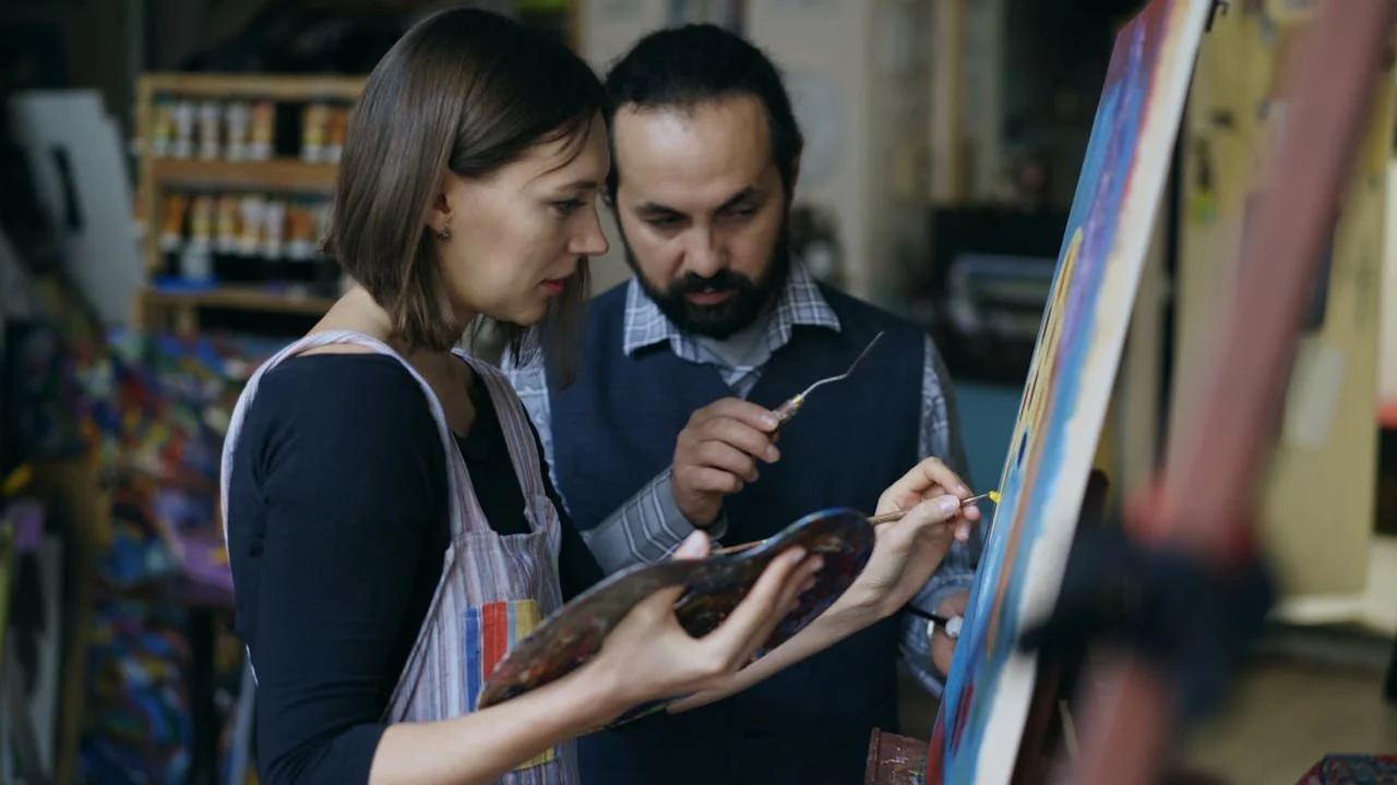 A teacher guides a student in an art class as she paints on a canvas