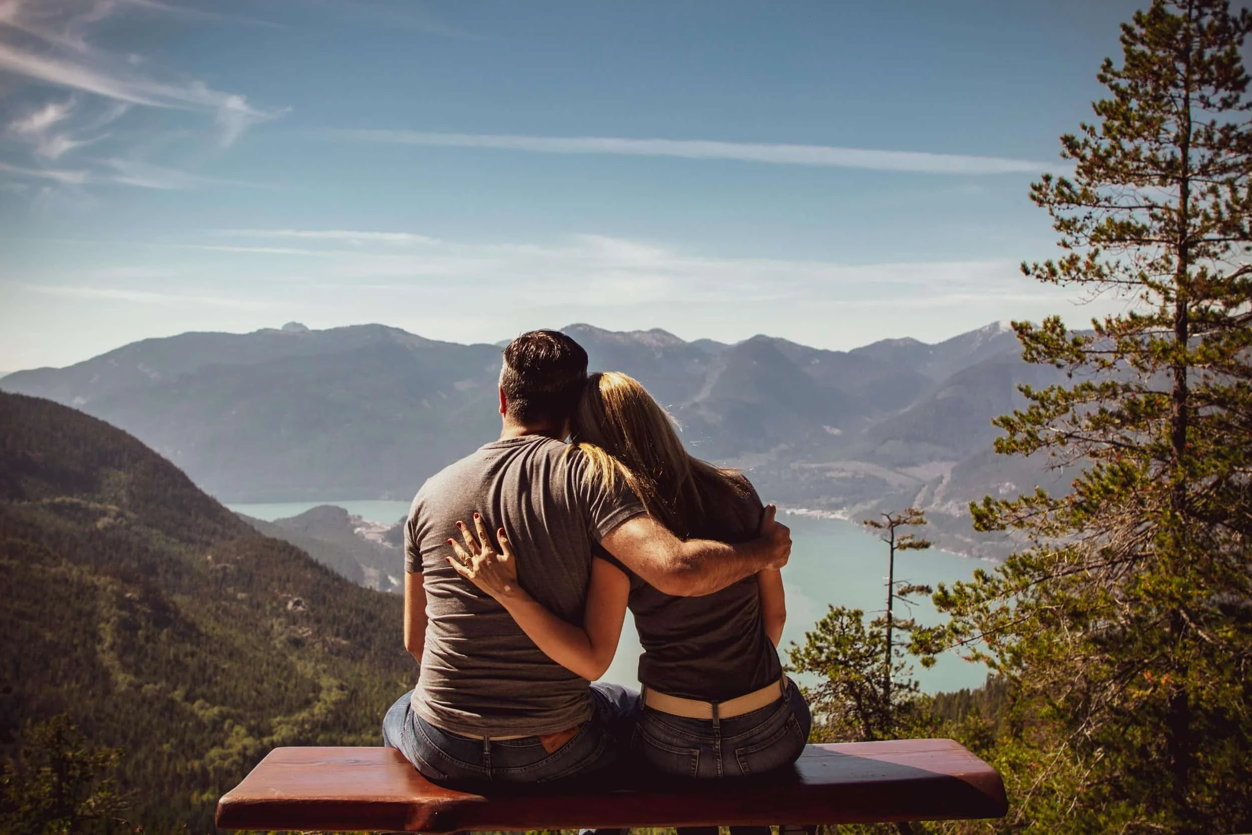 Couple sitting on a bench hugging, overlooking a mountain lake and pine trees under a partly cloudy sky.