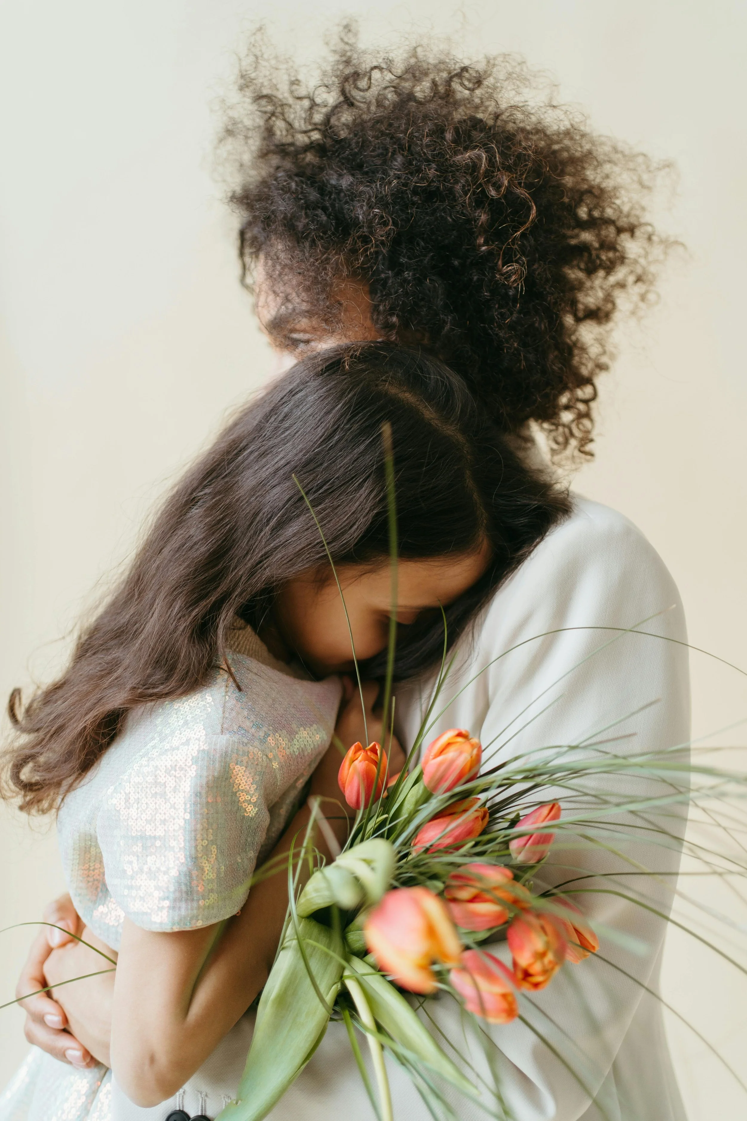 Two women, one with dark skin and curly hair wearing a white bandana, and the other with light skin and red hair with a pink headband, are smiling and embracing in a room with houseplants and white flowers.