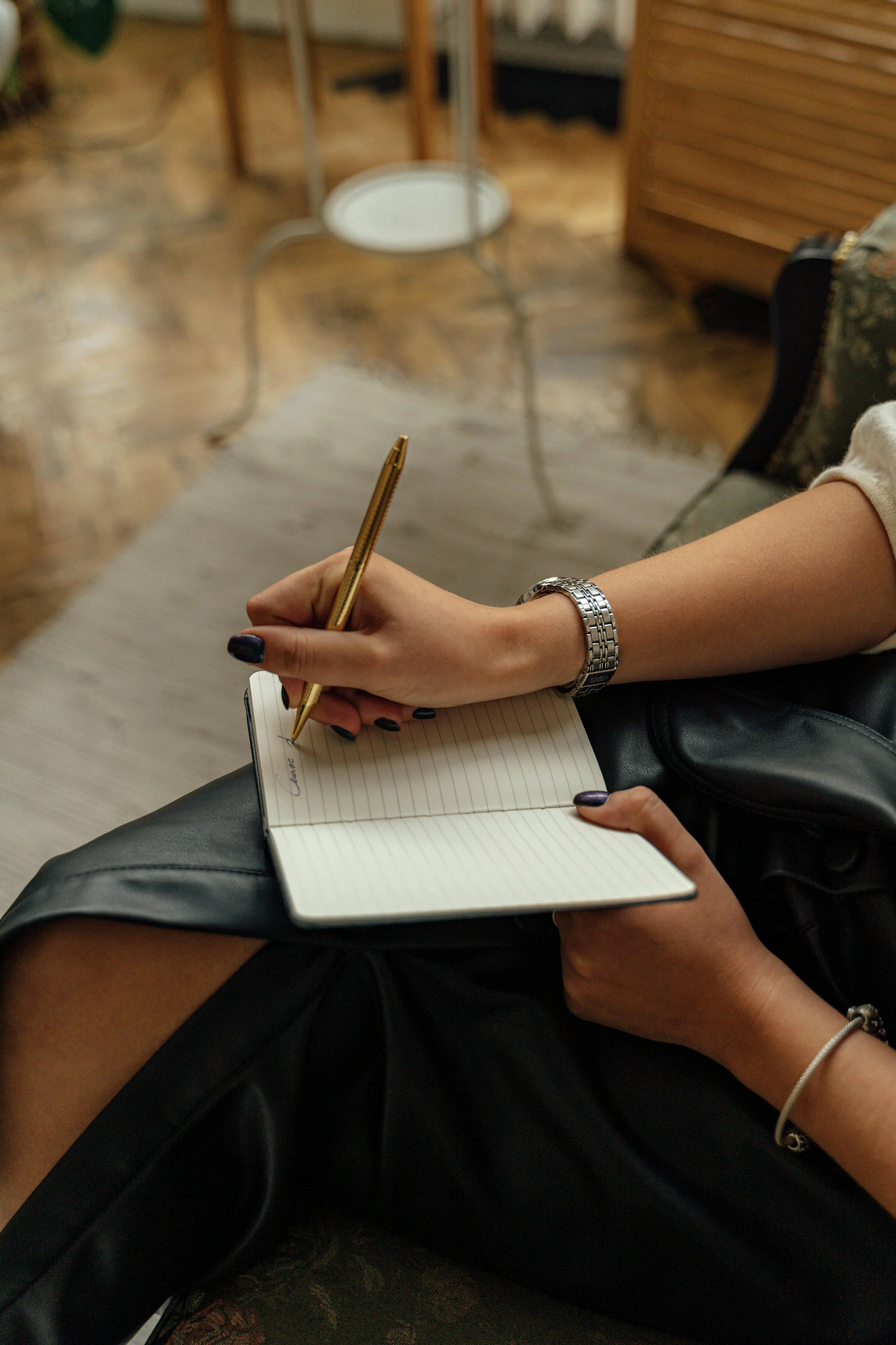 A person sitting on a black leather chair, writing in a lined notebook with a gold pen. They are wearing a silver watch and have painted nails. Another person is sitting above, with their legs resting on the lap of the person writing, wearing black clothing and a bracelet.
