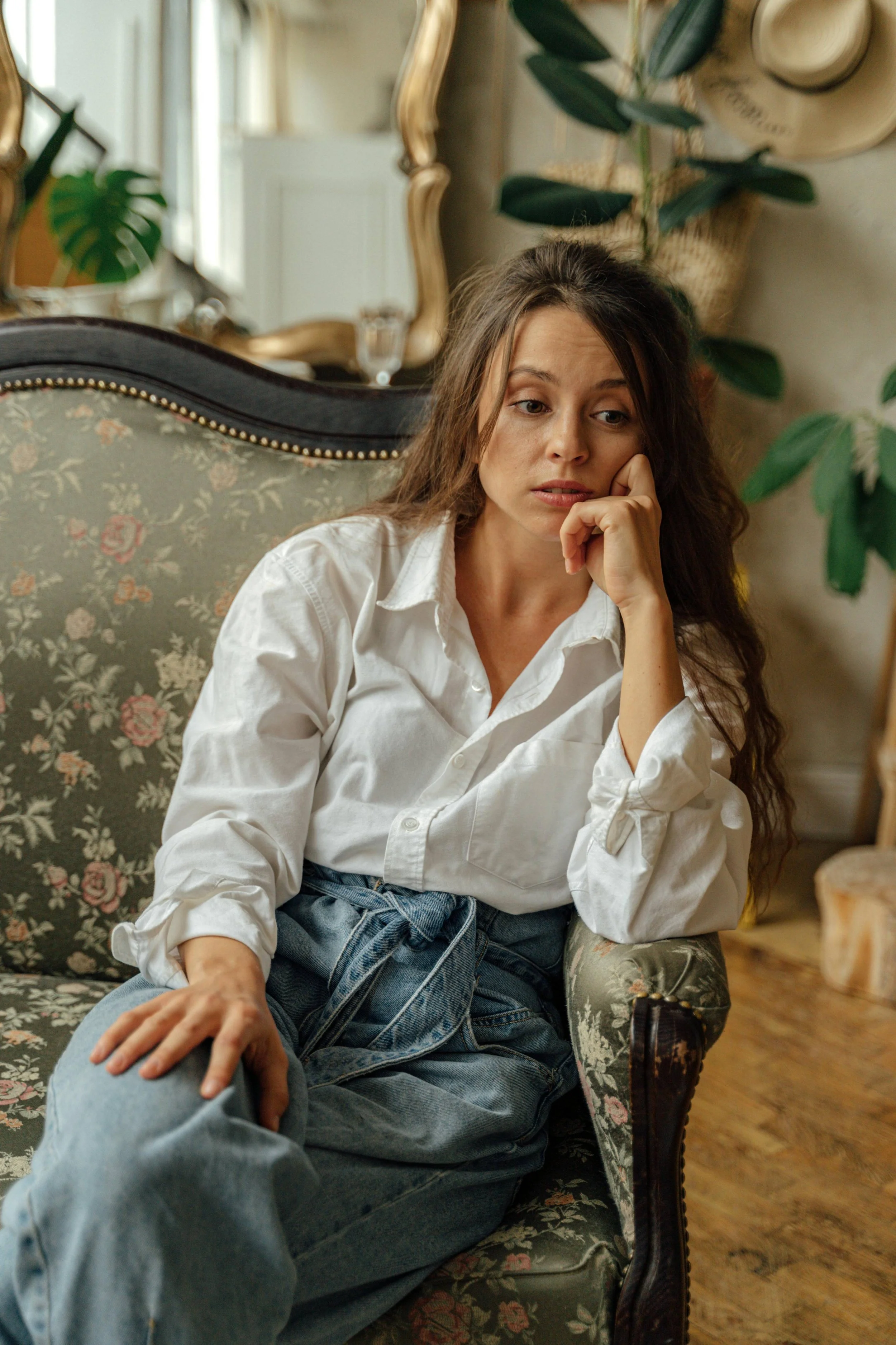 A woman with long, wavy brown hair, wearing a white shirt and blue jeans, sitting on a floral armchair in a cozy, decorated room, resting her chin on her hand with a thoughtful expression.