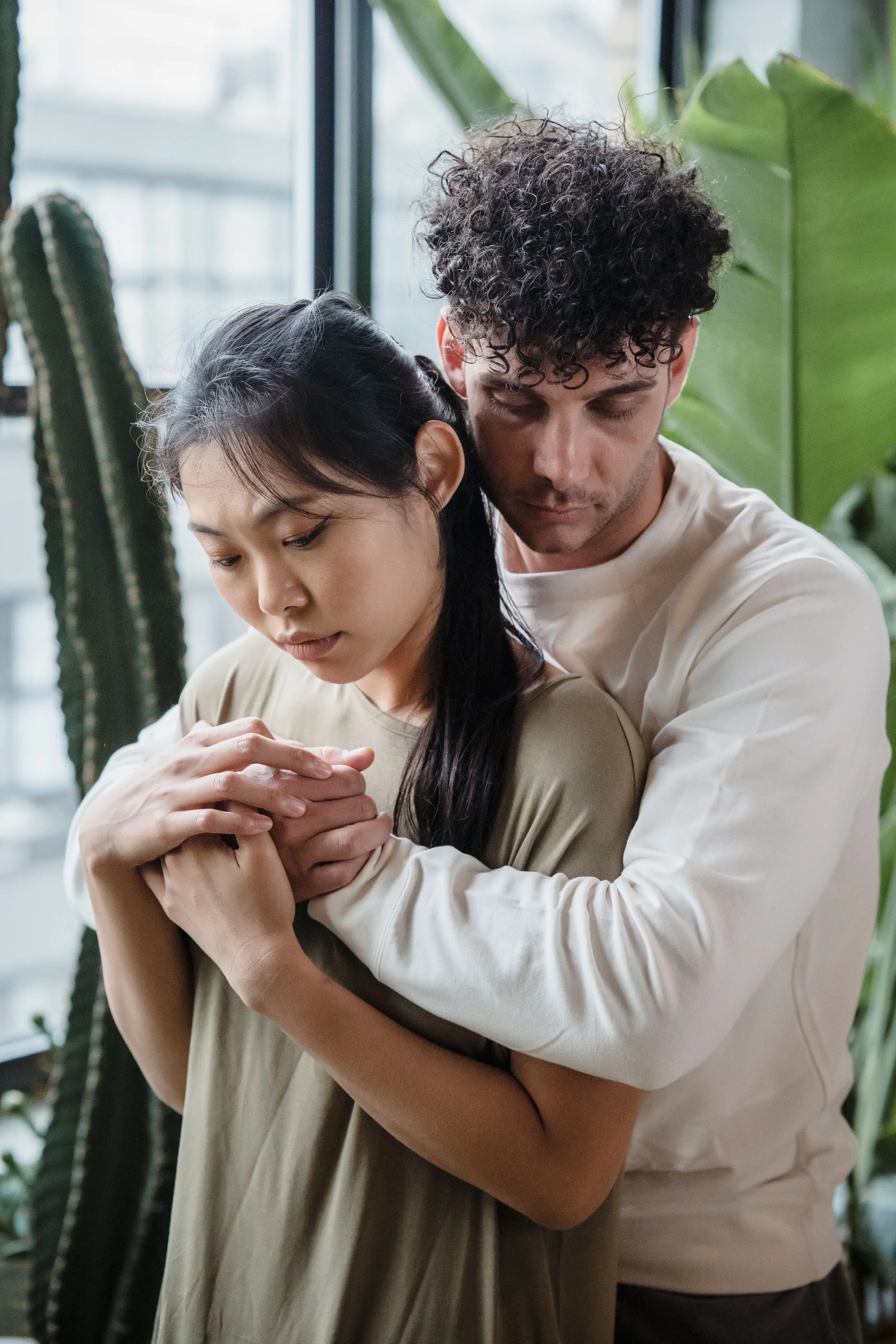 A man and woman embracing each other inside a room with large green plants and a window in the background.