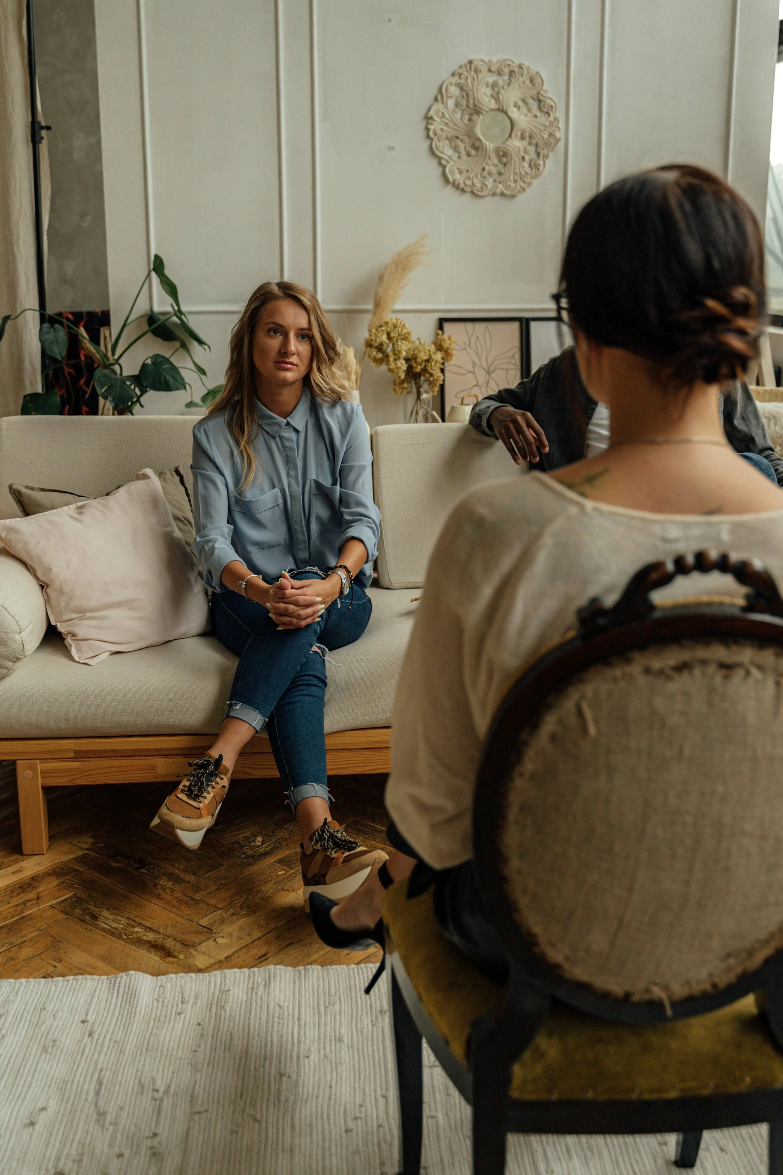 A woman with blonde hair sitting on a beige couch, talking to another woman with short dark hair and glasses in a patterned top, in a cozy, decorated room with plants and framed art