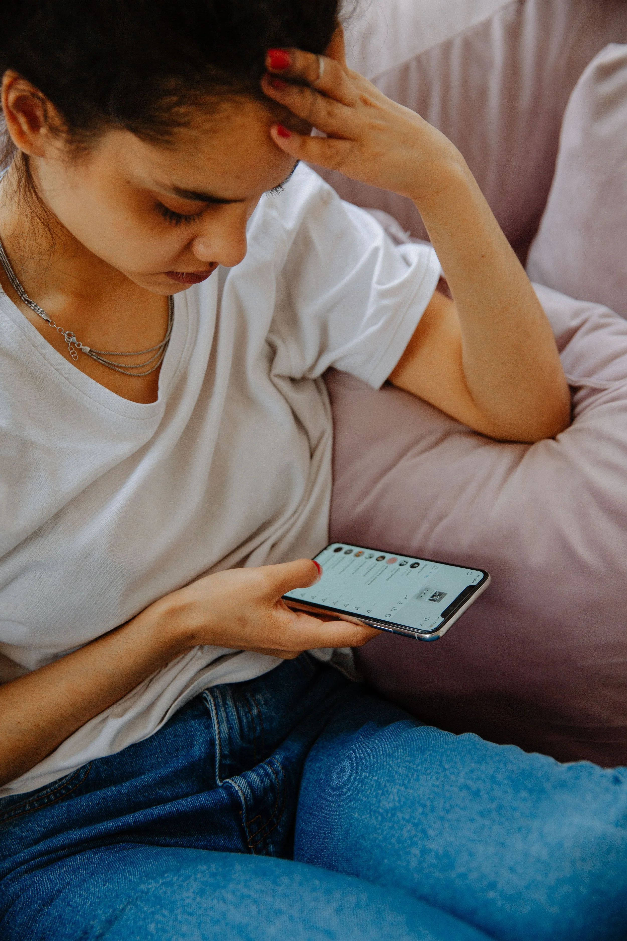 A young woman with red painted nails sitting on a bed, holding a smartphone, with her head resting on her hand.