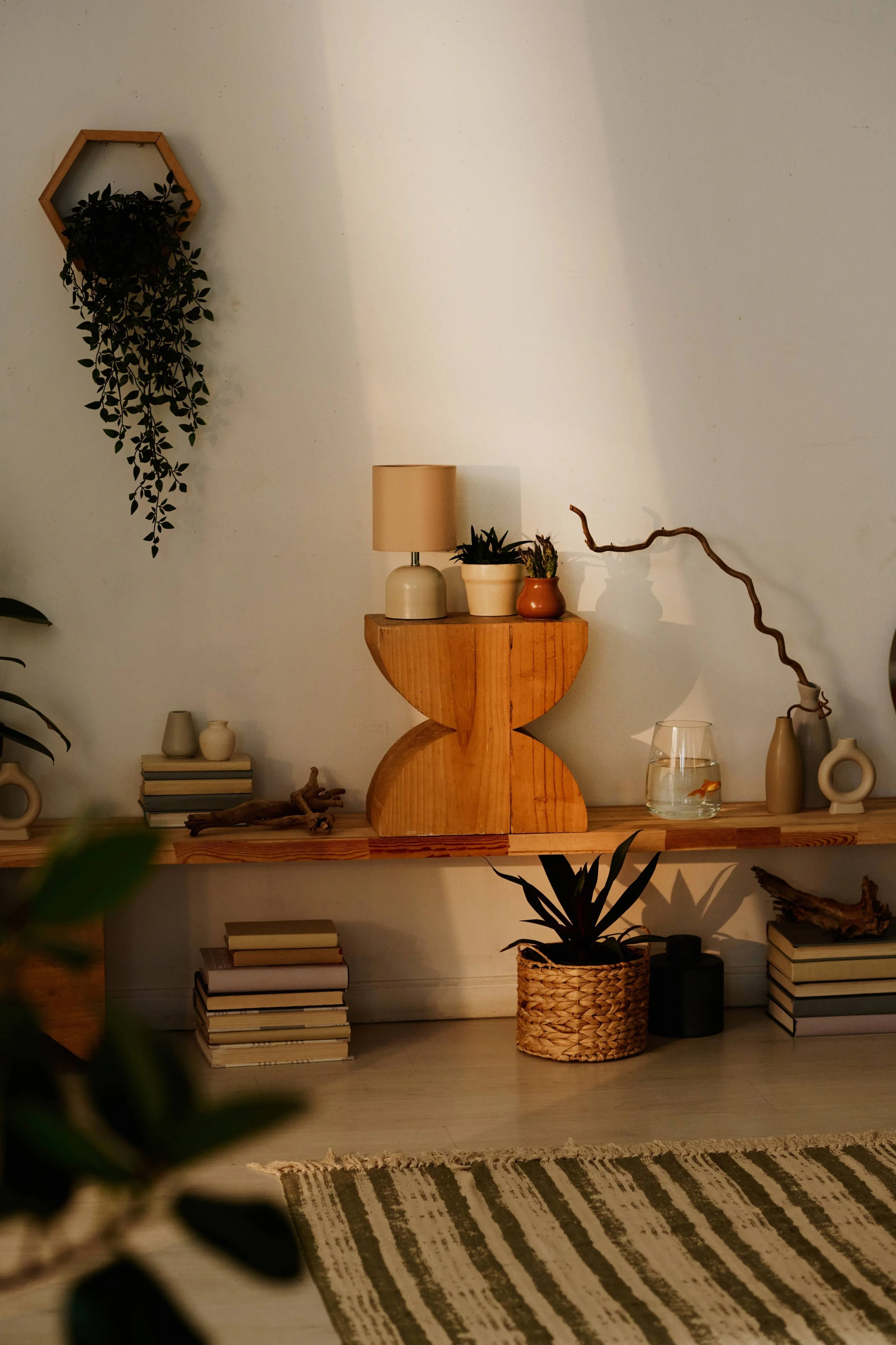 Interior decor with wooden shelves, potted plants, a lamp, stacked books, vases, and a striped rug.