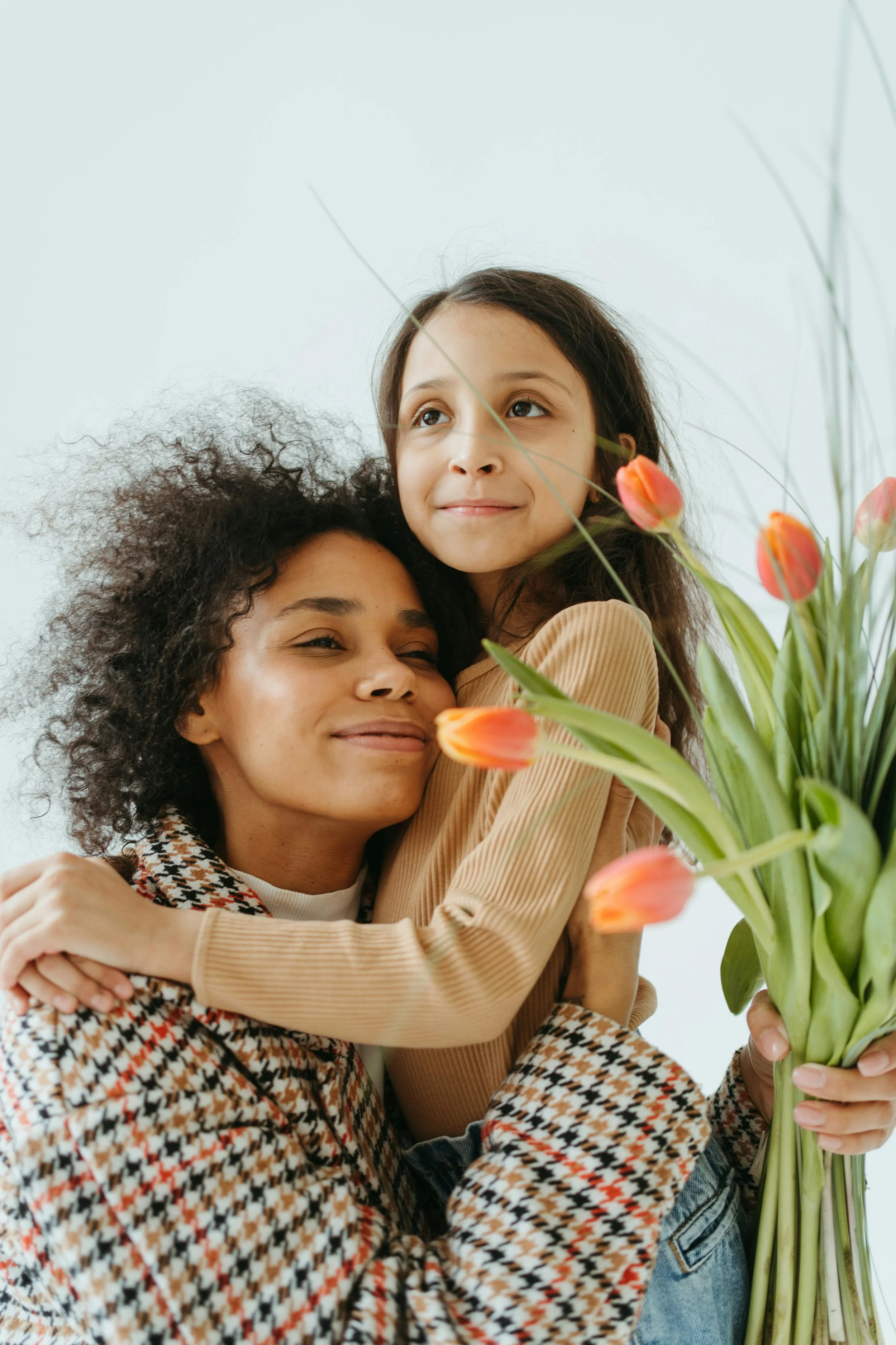 A woman and a girl sharing a hug, the woman is holding a bouquet of orange tulips, both smiling. The girl is sitting on the woman’s shoulders, and they are close to each other against a plain light-colored background.