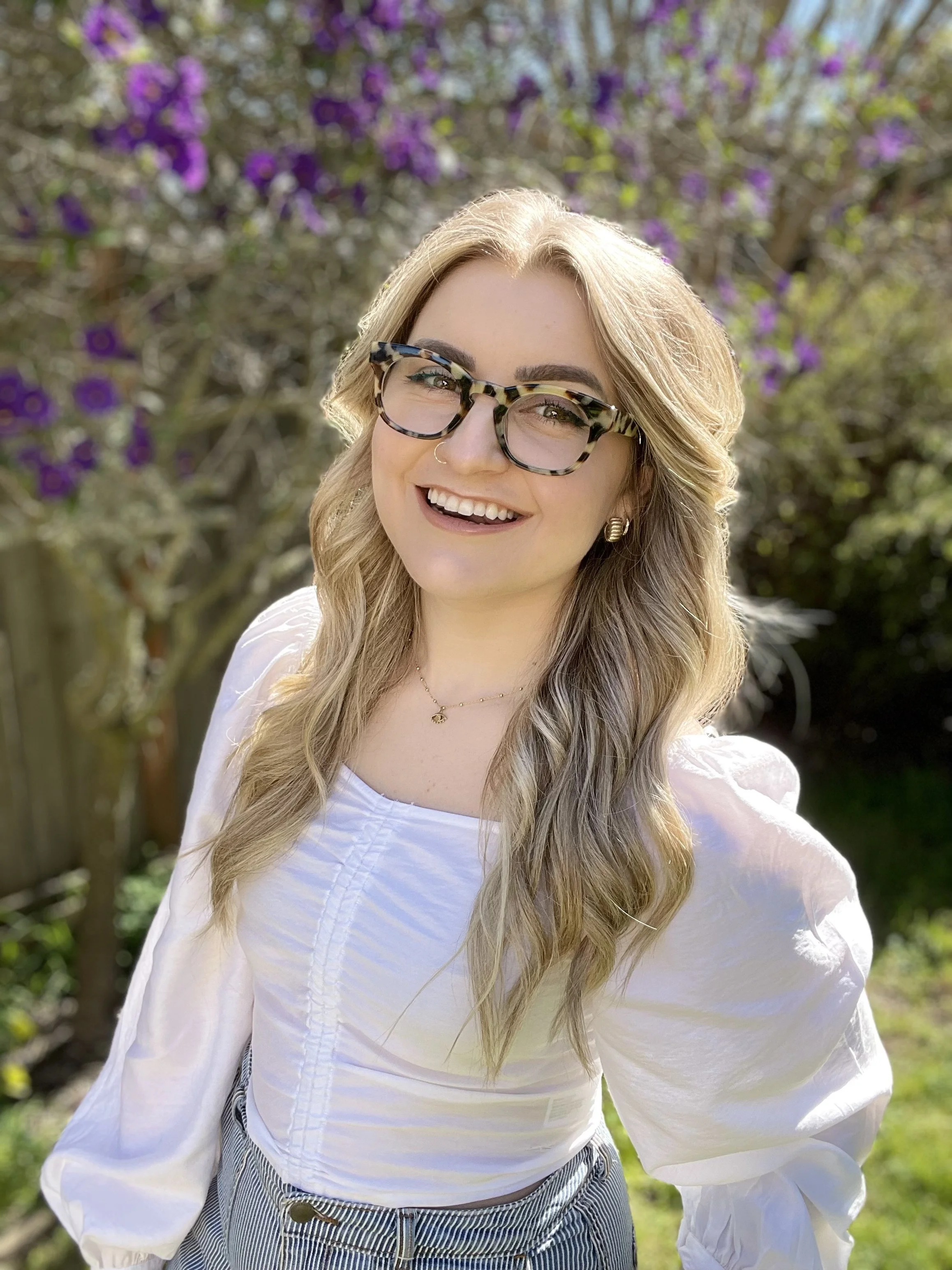 A young woman with blonde hair, wearing patterned glasses, a white blouse, and striped pants, smiling outdoors with purple flowering trees in the background.