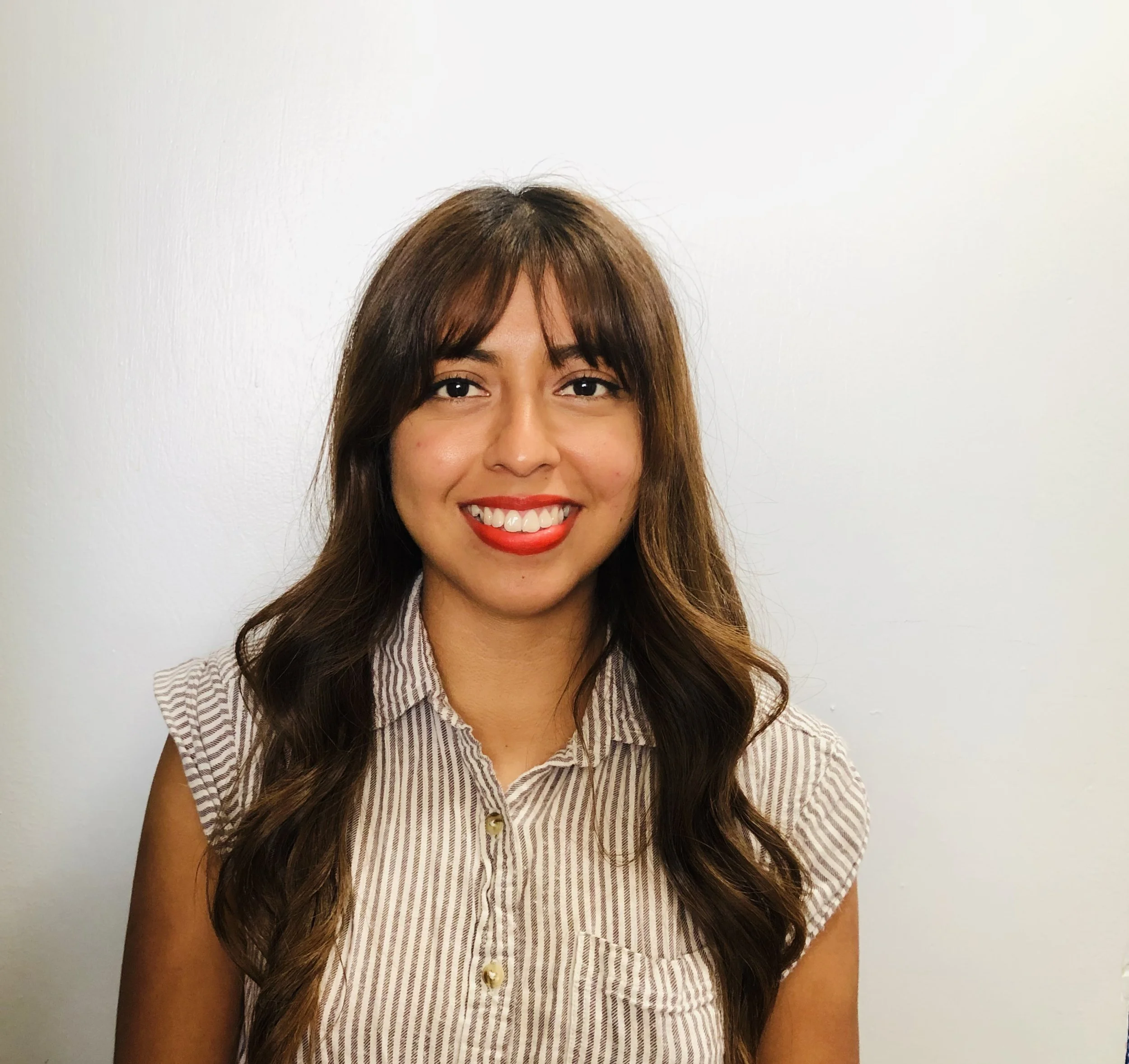 A young woman with long wavy brown hair, smiling with red lipstick, wearing a light-colored, striped blouse with beige buttons, standing against a plain white wall.