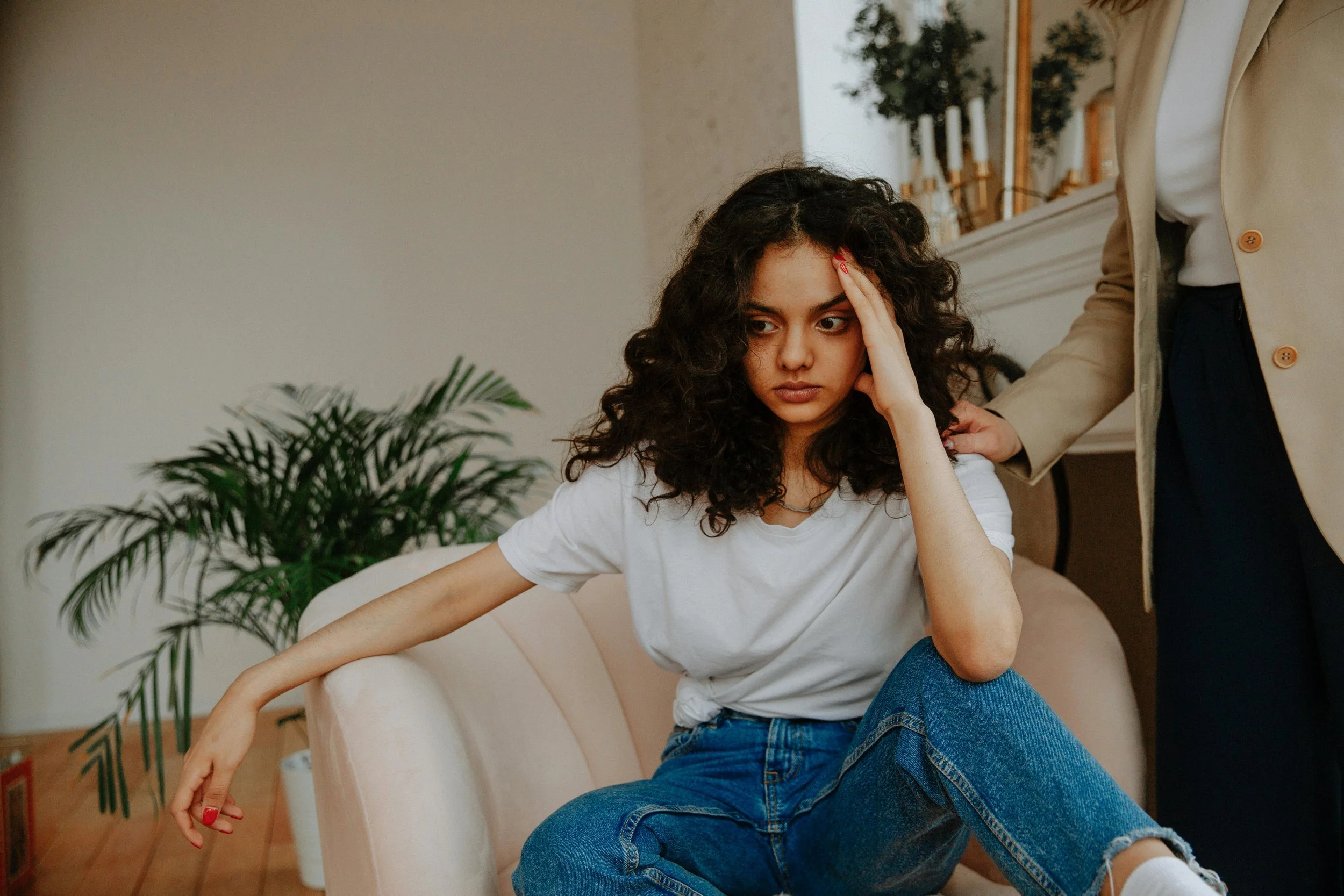 A woman with curly dark hair sitting on a couch, holding her forehead with her hand, appearing distressed. She is wearing a white shirt and blue jeans. An older person in a tan coat is standing beside her, with their hand on her back. In the background, there are candles on a mantel and a large mirror, with a green plant nearby.