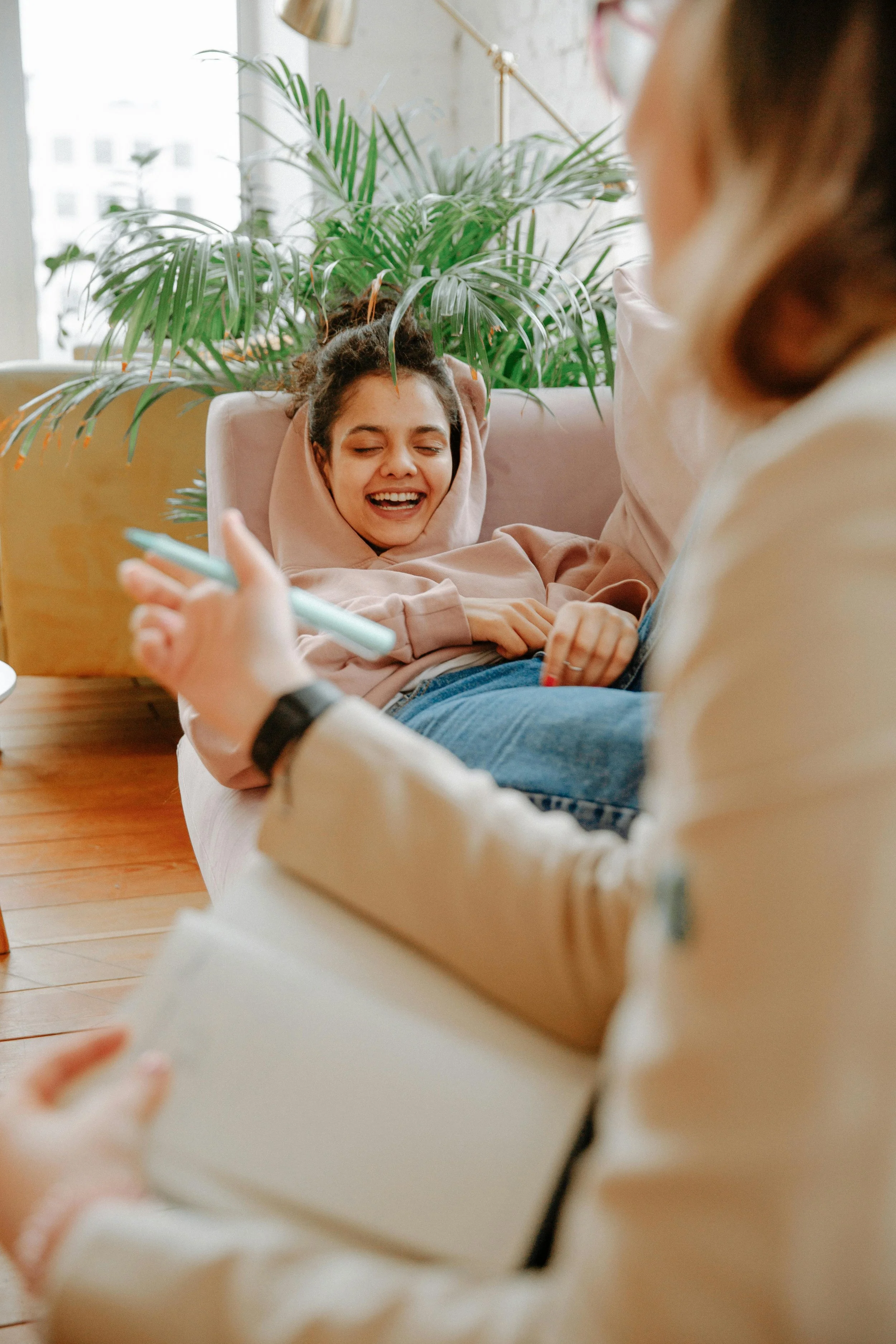 A woman with dark curly hair, wearing a pink hoodie, is lying on a pink couch, smiling with her eyes closed. She is next to a large green plant and is interacting with two other people, one holding a pen and the other partially visible.