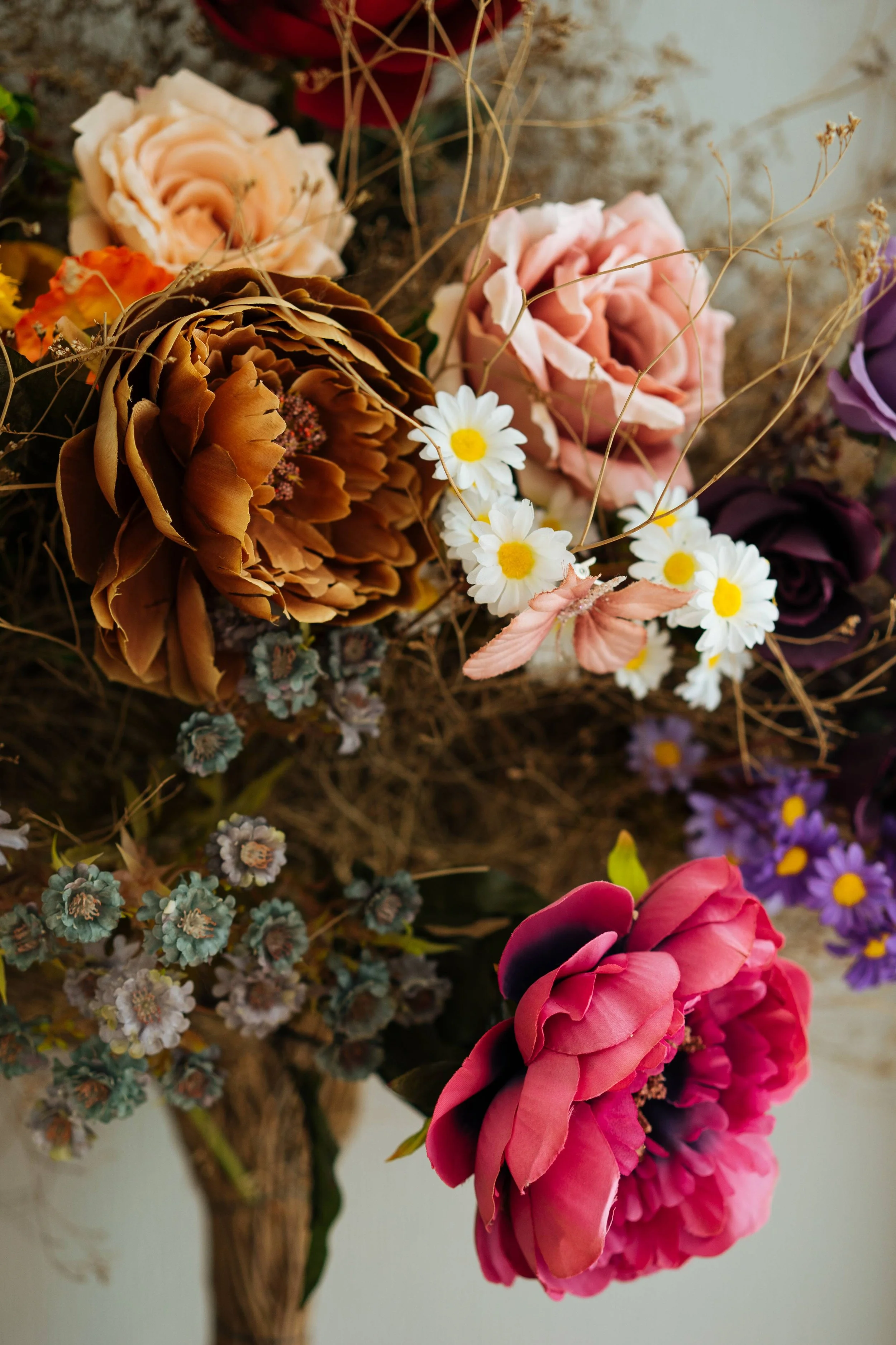 A colorful bouquet of various flowers, including a large pink peony, white daisies, a brown peony, and purple, yellow, and pink flowers, arranged with dried branches.