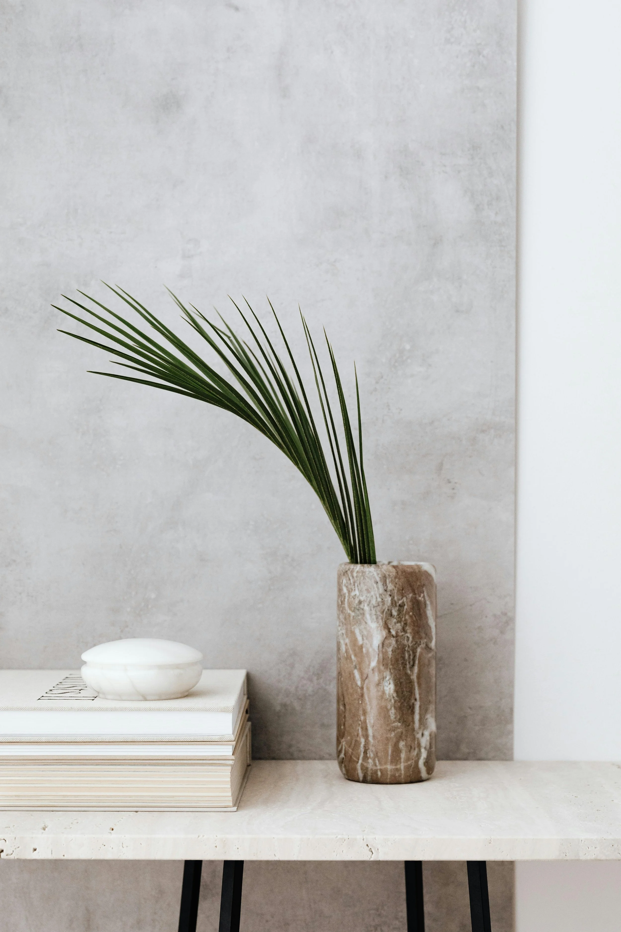 A minimalist decor setup featuring a potted palm branch in a marble vase, a white ceramic dish on a stack of books, all on a white table against a textured wall.